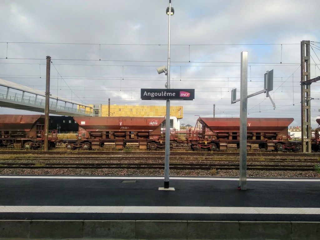 View of Angoulême train station, showing the platform, railway tracks, freight cars, overhead power lines, a sign with the station's name, and a cloudy sky.