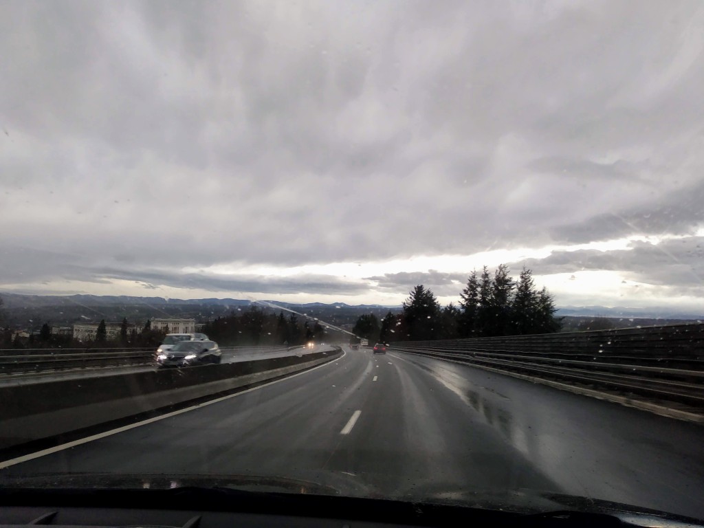 View from inside a car driving on a wet motorway under a cloudy, overcast sky.  A dark car drives in the left lane; the asphalt reflects light from the rain. In the distance, a line of trees and low buildings is visible against the horizon, a faint mountain range beneath the heavy cloud cover.