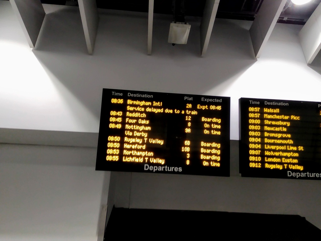 Train station departure board with times, destinations, platform numbers, and expected departure status, mounted on a white wall under white ceiling beams. The 08:36 to Birmingham International reads 'Service delayed due to a train'.
