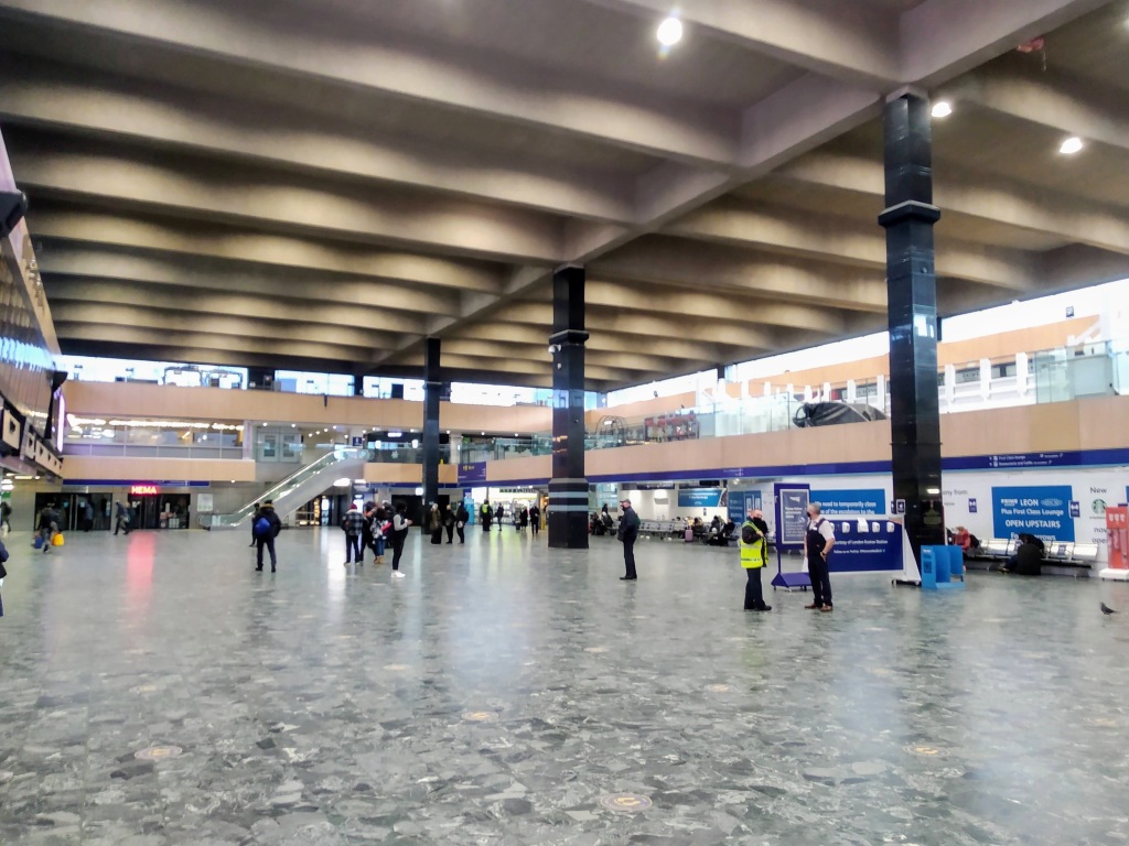 Interior of the station concourse at London Euston, with high ceilings, marble floors, and several black structural pillars. People are sparsely distributed throughout the space.