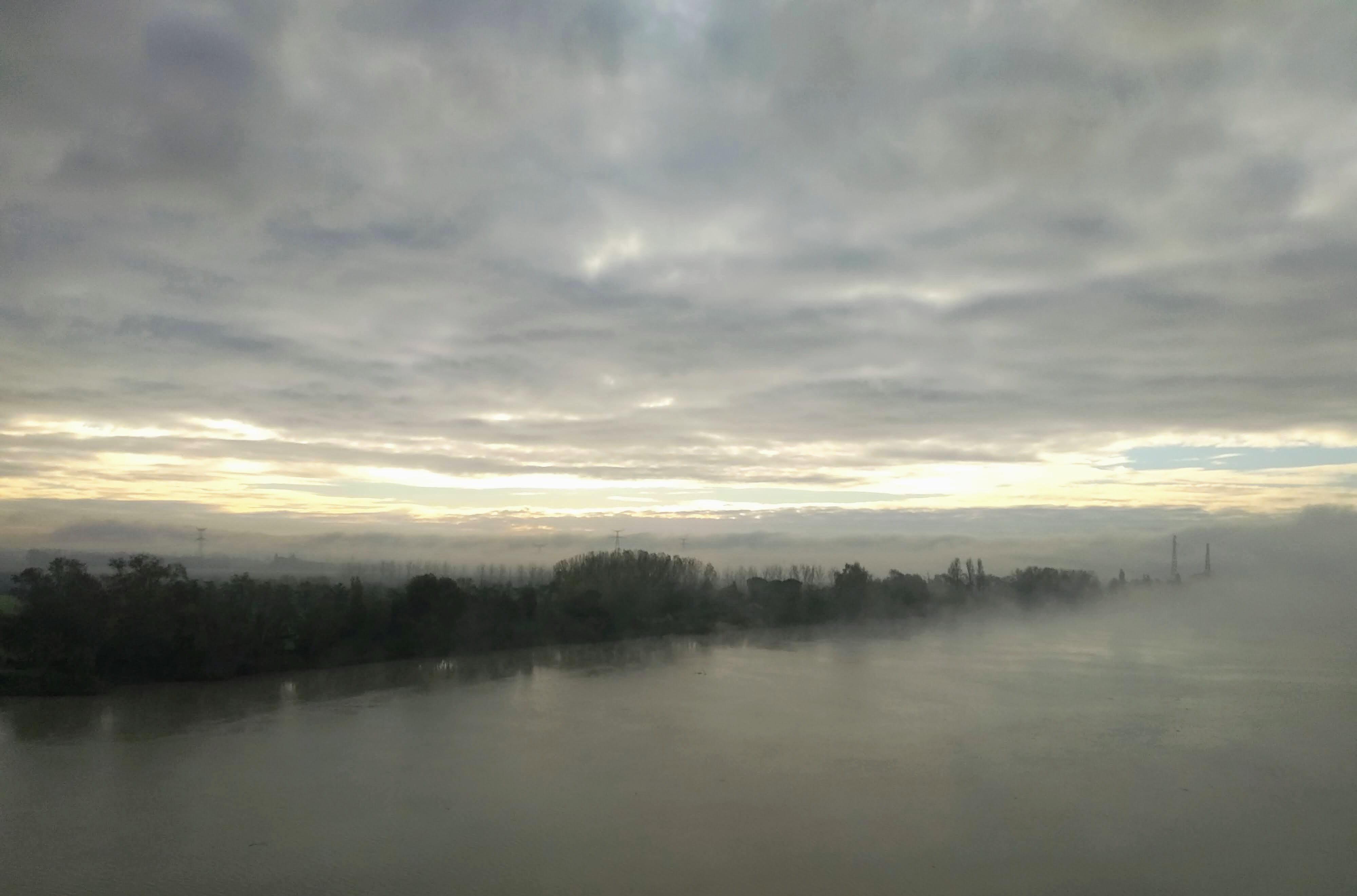 Foggy landscape view of a wide, flat river with a tree-lined bank, under a heavily clouded, overcast sky with hints of the sunrise visible at the horizon. Silhouetted trees are partially obscured by low-lying fog or mist that blankets the water.