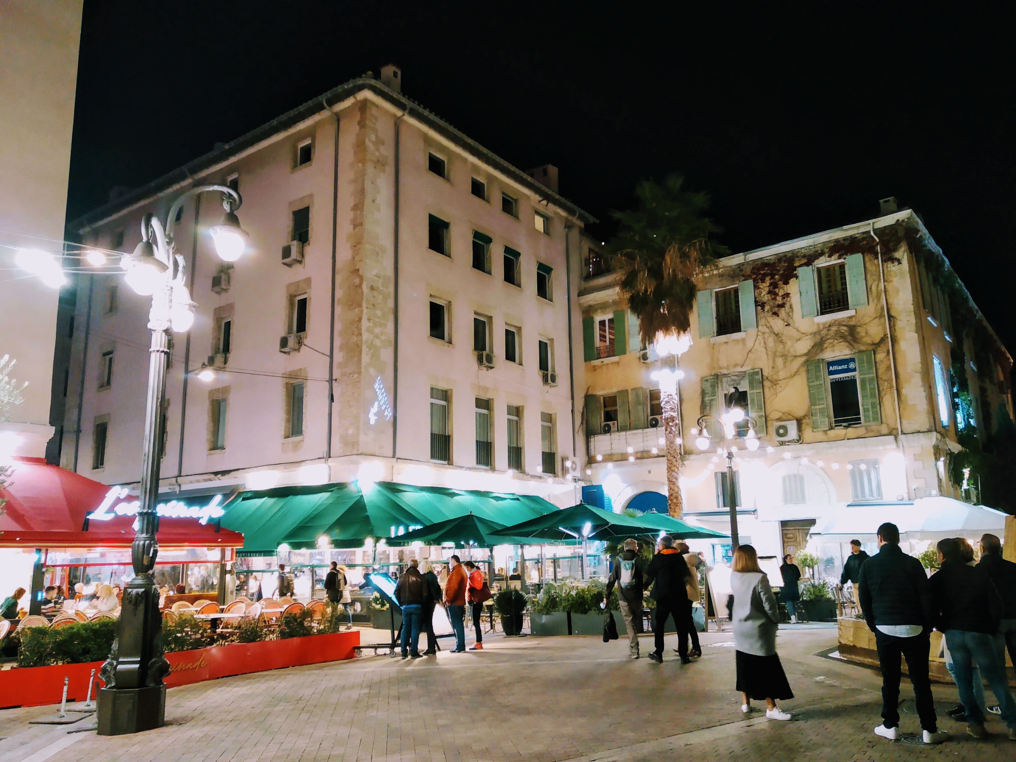 Nighttime view of a European city square with people gathered around outdoor restaurant seating under green awnings, lit by streetlights and building lights, with pastel architecture and a palm tree.
