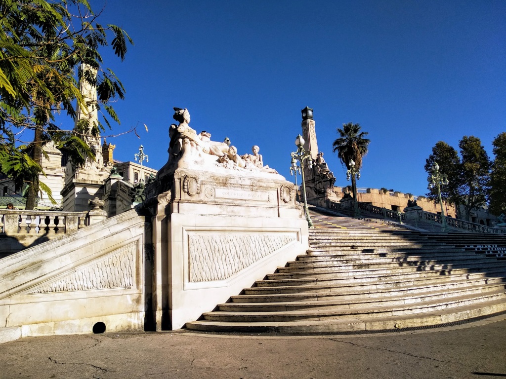 A grand, tiered white stone staircase leads up to a monumental sculpture featuring reclining figures, set against a backdrop of clear, bright blue sky and palm trees, showcasing classical architecture and a sunny, inviting ambiance.
