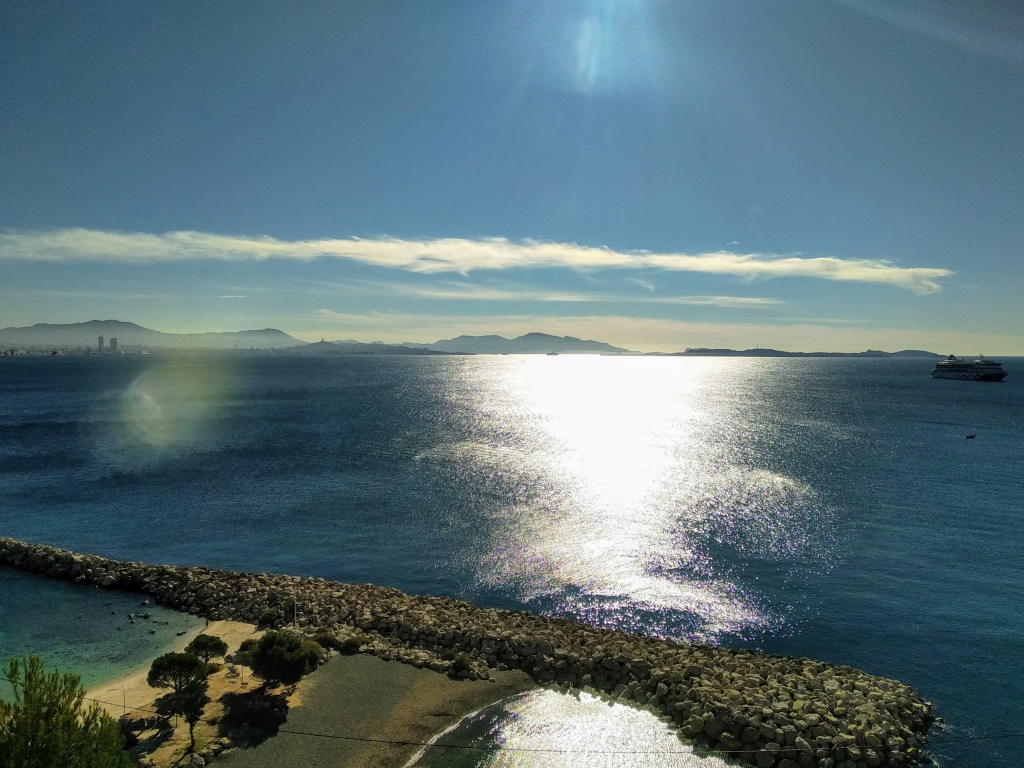 Bright sunlight reflecting on a vast blue ocean, partially obscured by light clouds, viewed from an elevated perspective with a rocky breakwater in the foreground and a distant cityscape and mountains on the horizon. A cruise ship is seen on the sea.
