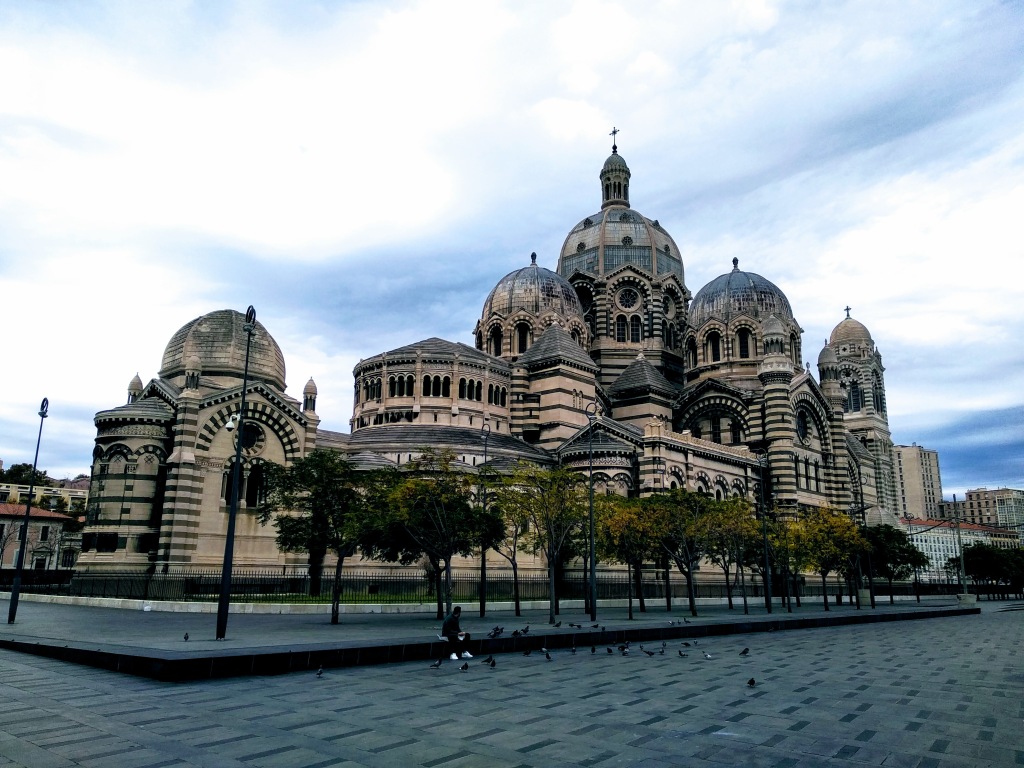 View from a plaza of the Roman-Byzantine style Marseille Cathedral, a large stone building with striped patterns on its domes and walls, under a cloudy sky.