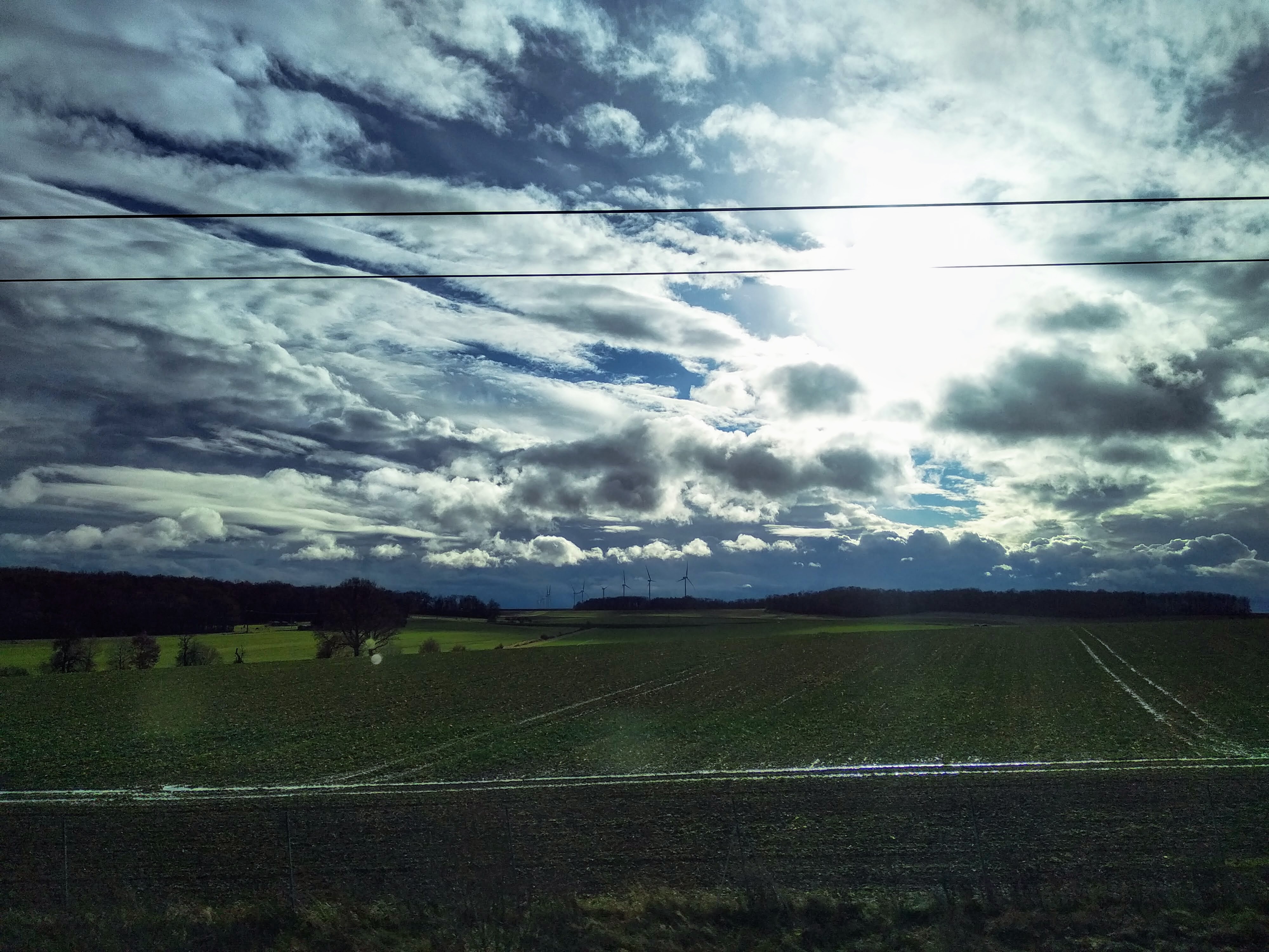 Dramatic sky with scattered, puffy grey and white clouds and patches of blue above a green, cultivated field. The sun shines brightly behind clouds in the centre. Wind turbines are faintly visible on the horizon.