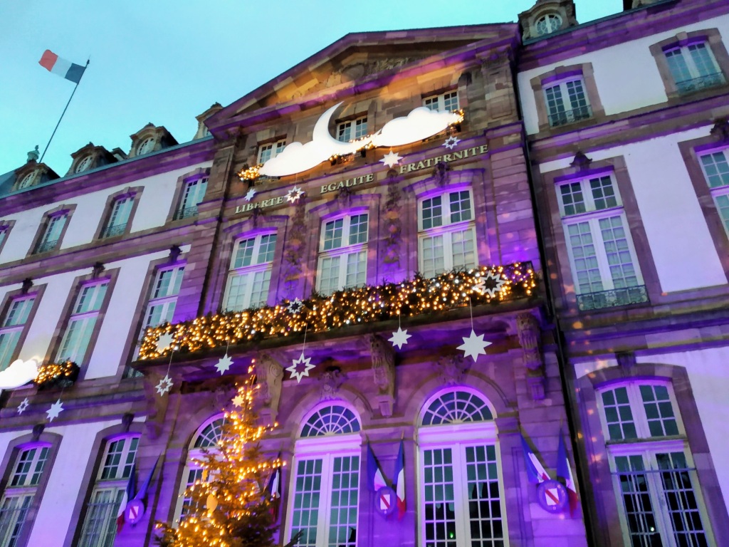 Illuminated historic French building with "Liberté, Égalité, Fraternité" inscription, adorned with Christmas decorations including lights, star ornaments, and a small illuminated Christmas tree in front, under a twilight sky, with a French flag on top.