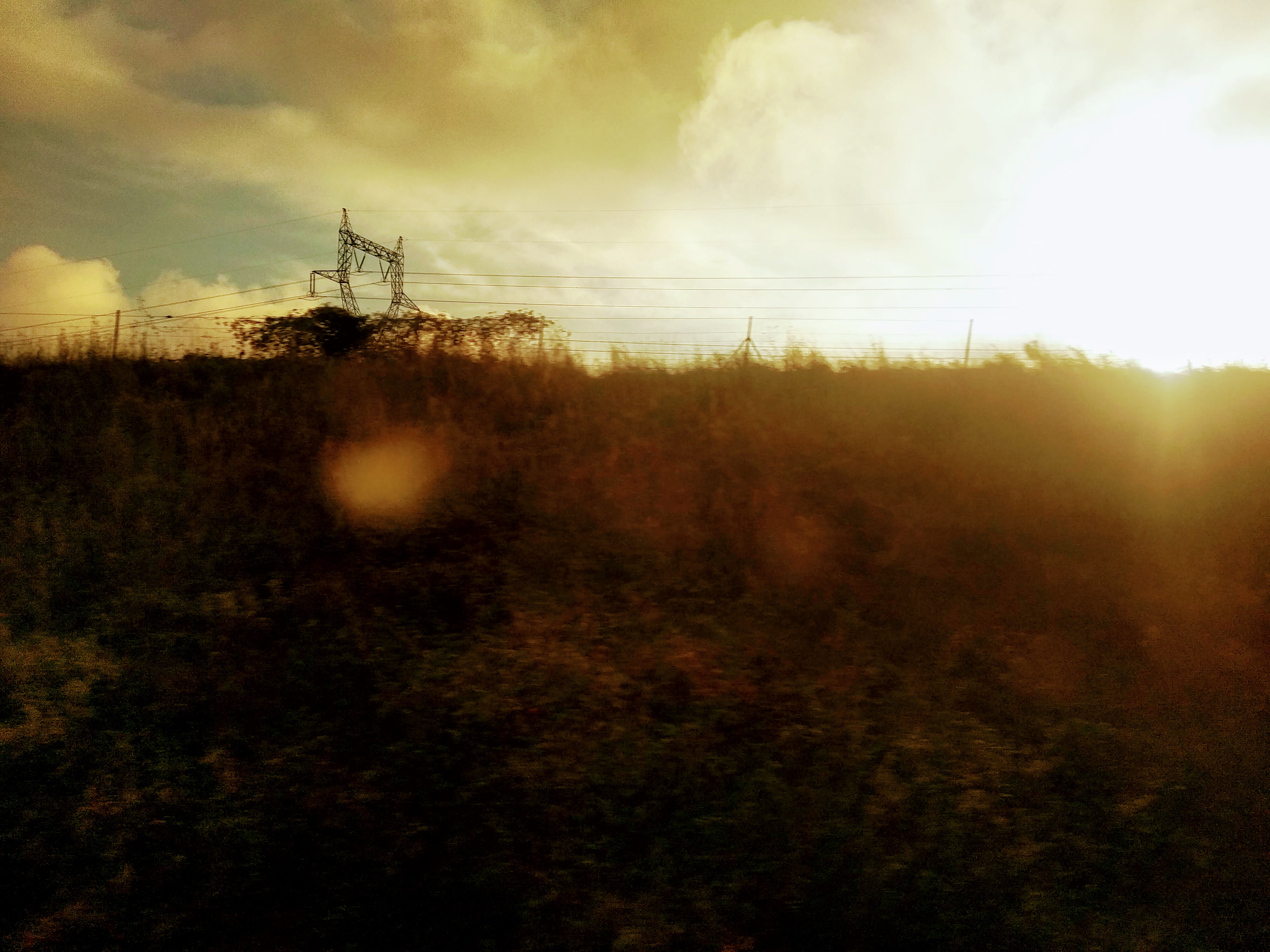 Silhouette of a high voltage power pylon against a cloudy sky, back-lit by the rising sun over a grassy hillside.