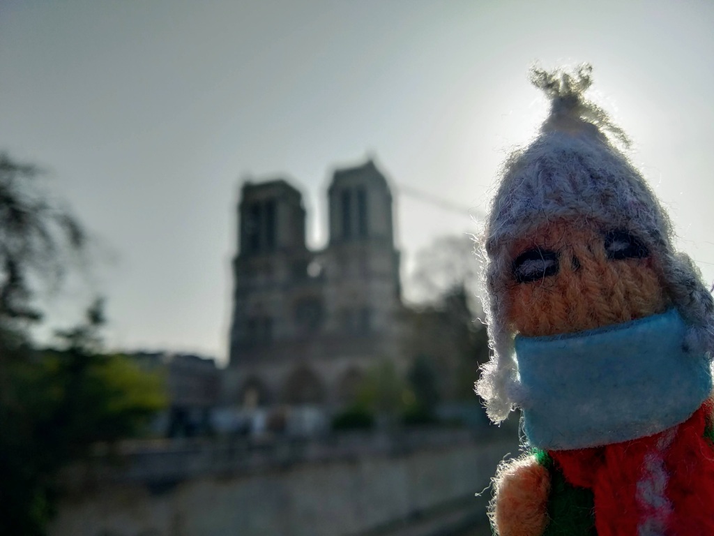 Close-up of a knitted doll wearing a surgical mask in front of the Notre Dame cathedral in Paris, France, on a sunny day.
