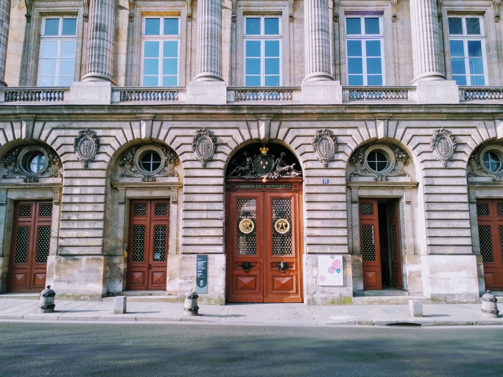 Facade of a classical style building with a series of arched entrances, carved stone ornamentation, columns, and arched windows, featuring a large central door with ornate brass fixtures and the inscription "ADMIN DES MONNAIES" above it.
