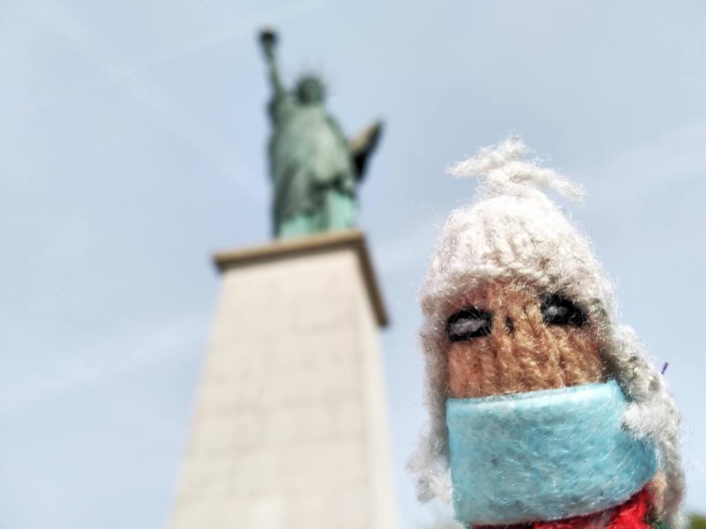 Close-up of a knitted finger puppet wearing a face mask, with a Statue of Liberty blurred in the background.