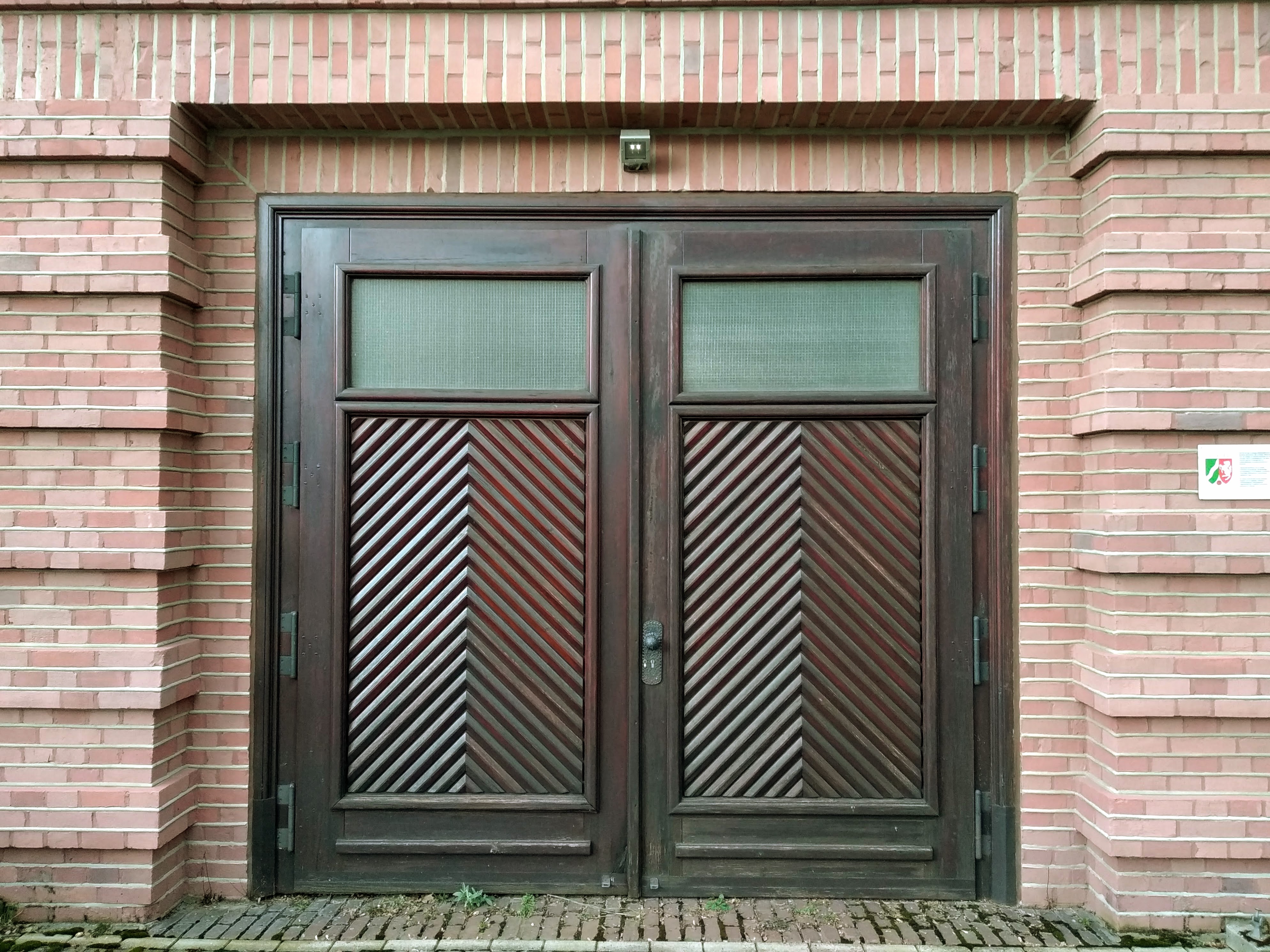 A brown wooden double door with dense chevrons in a red brick wall.