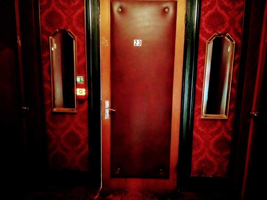 A deep red leather door numbered 23 flanked by red velour wallpaper and mirrors.