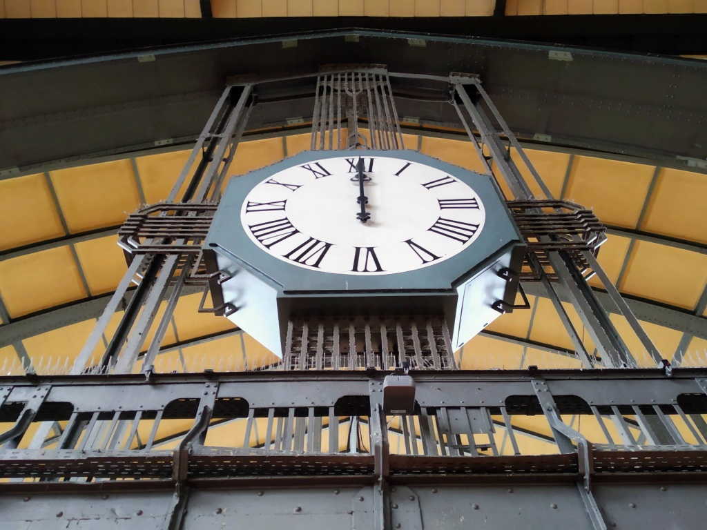 An octagonal steel clock displaying 12:00 under the roof of Hamburg Hauptbahnhof.