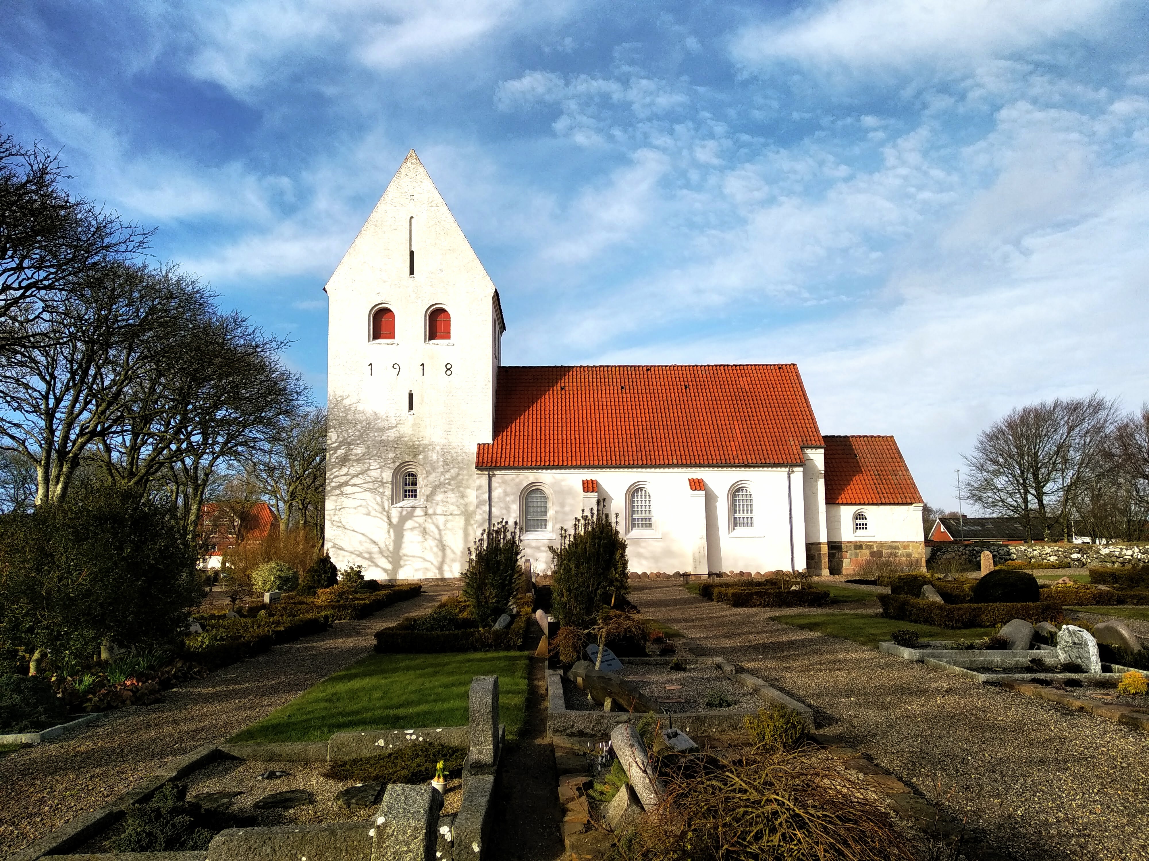 Jegindø Church, a white building with a red tile roof built in 1918,  seen across its graveyard.