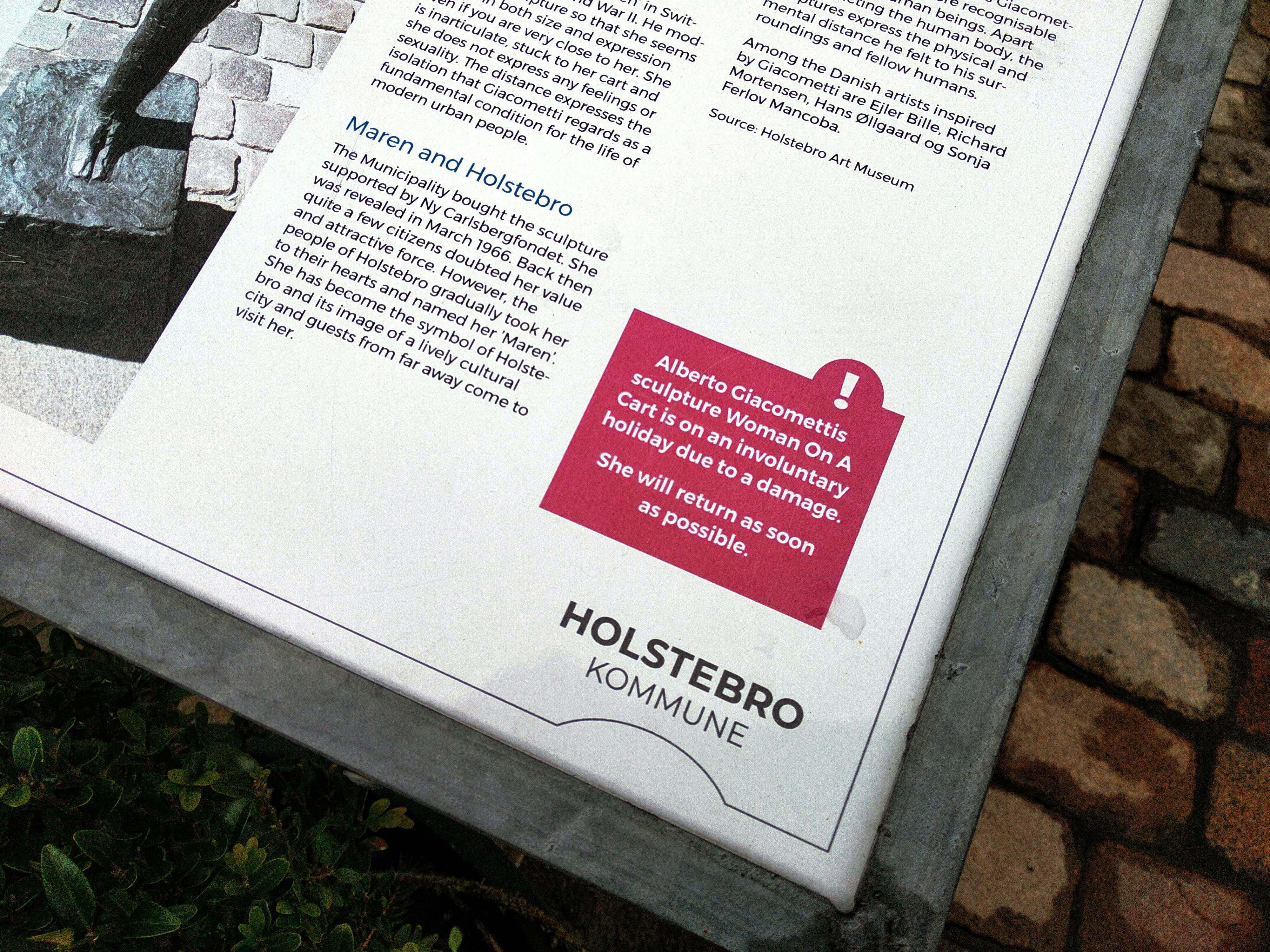 Text from a plinth reads: "Alberto Gicometti's sculpture 'Woman On A Cart' is on an unvoluntary holiday due to a damage. She will return as soon as possible."