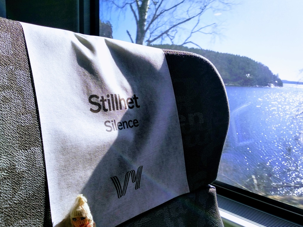 Puppet sitting in a railway seat with a lake passing the window. The antimacassar reads "Silence".