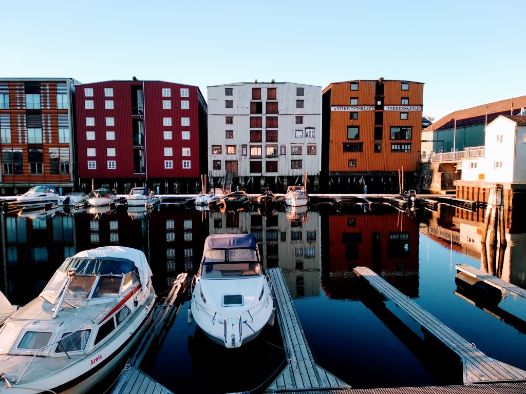 Boats moored on the canal in front of Trondheim Sentralstasjon in front of colourful wooden buildings on stilts. Blue skies.