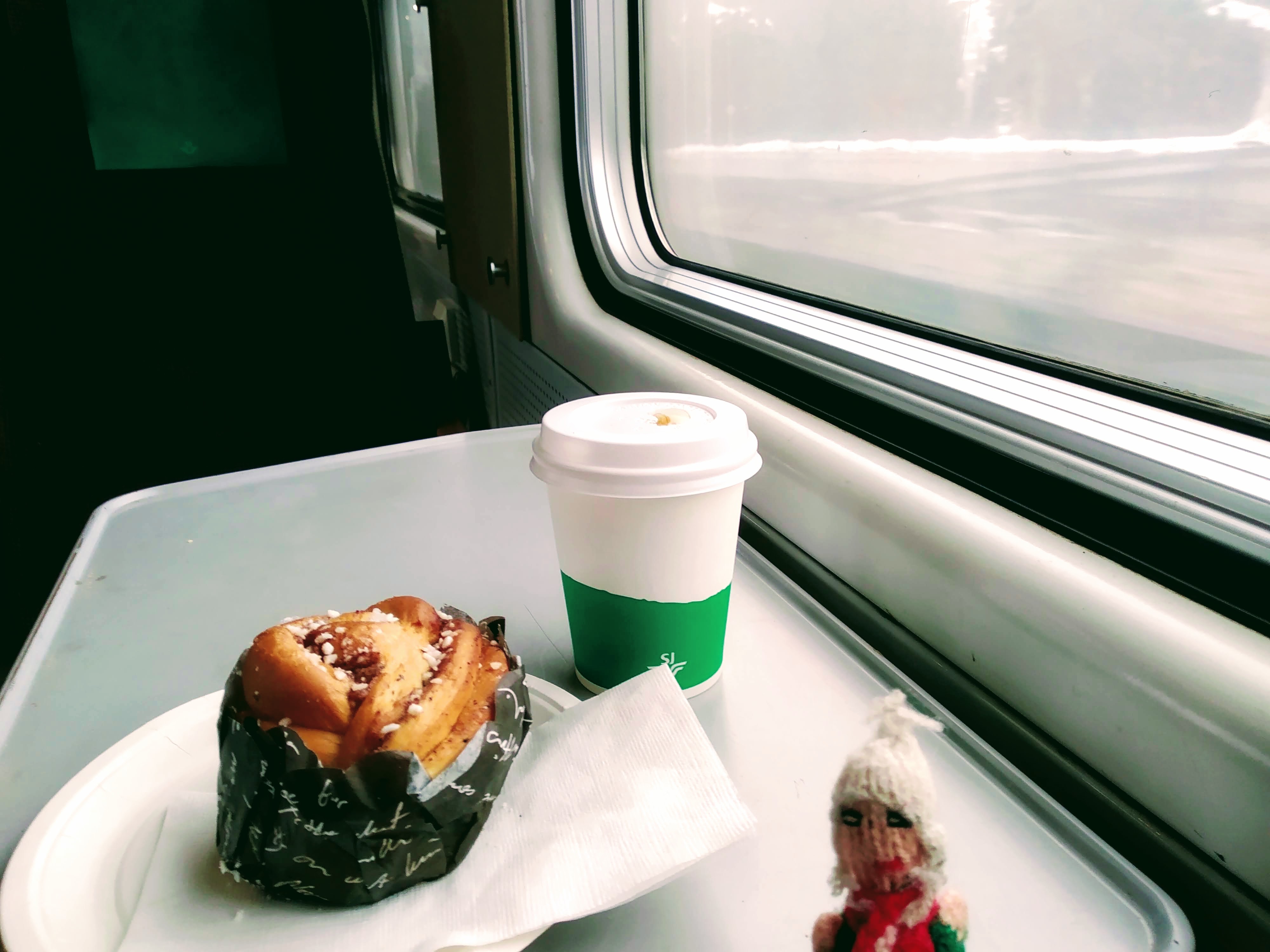 A cinnamon roll and a mug of coffee next to a window on an SJ Intercity train.