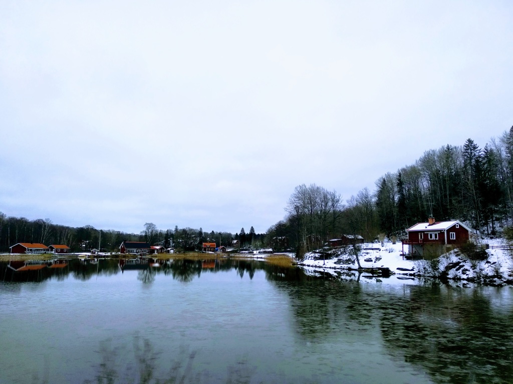 Lake-side houses covered in snow. The lake is clearly thawing. The sky is grey.