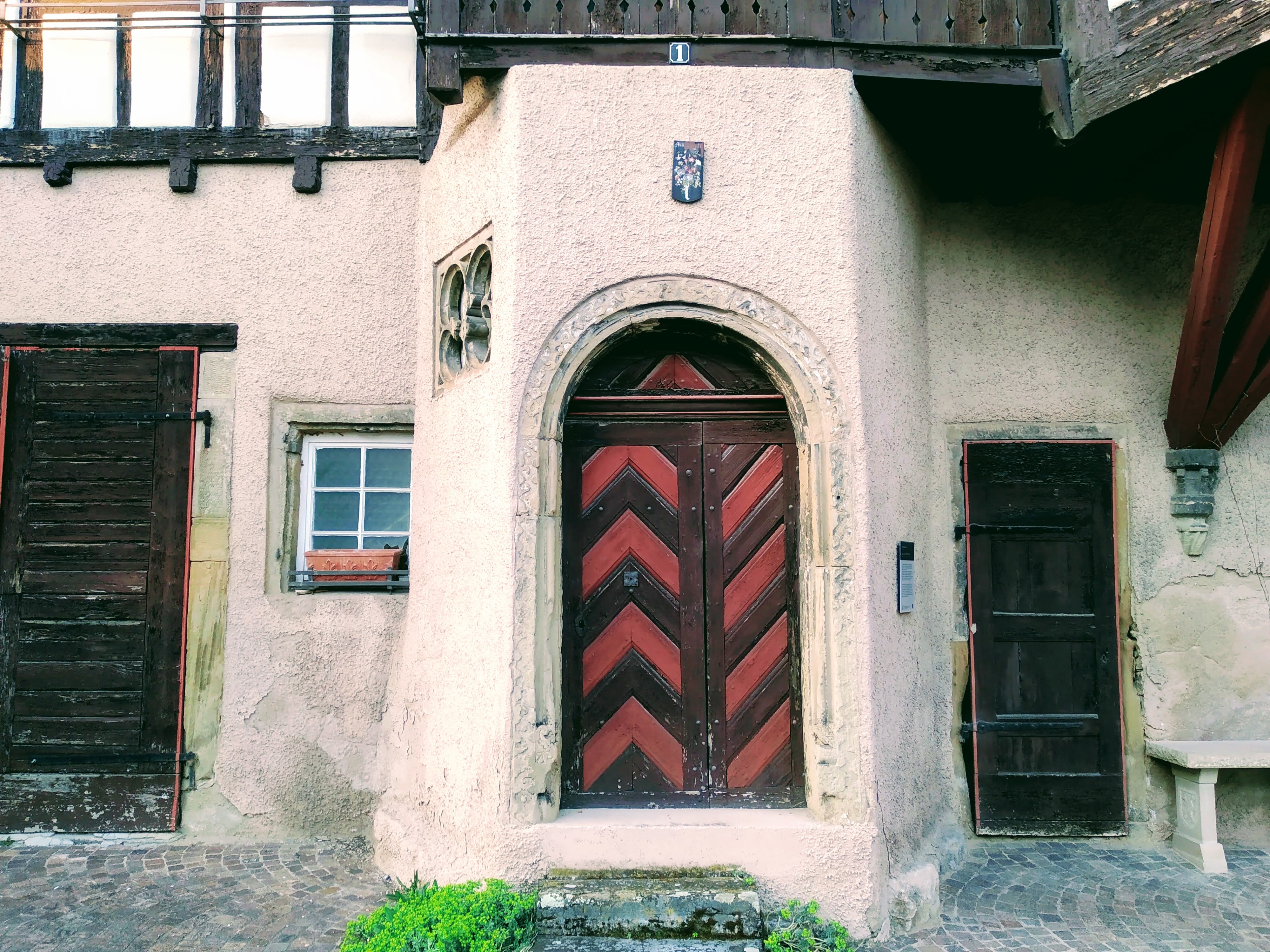 An old wooden door with red and brown chevrons, in a beige wall of a black and white house.