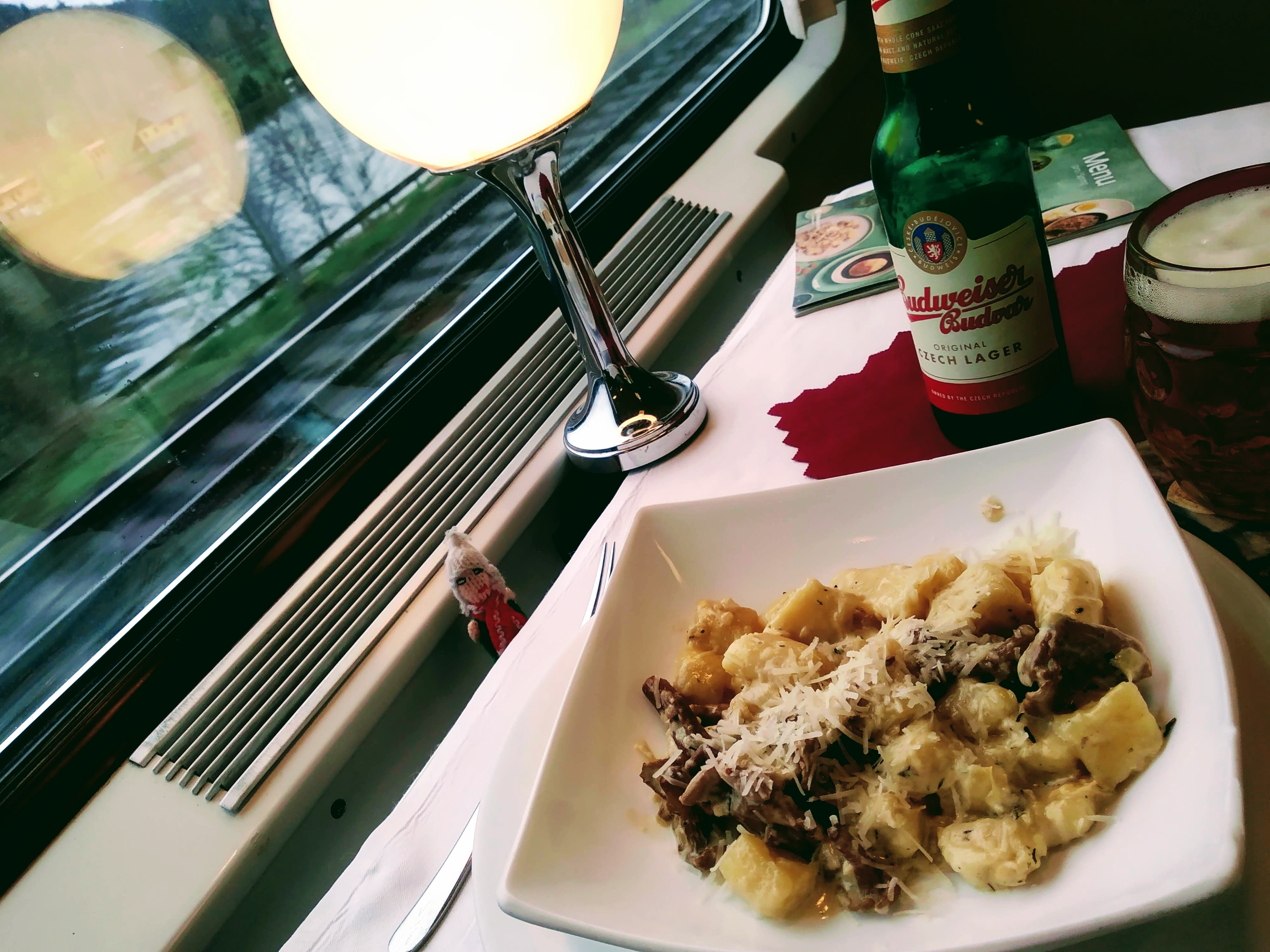 A picture of gnocchi in a china bowl accompanied by a glass of Budvar beer served on a table in a dining car. The river Elbe is visible through the window.