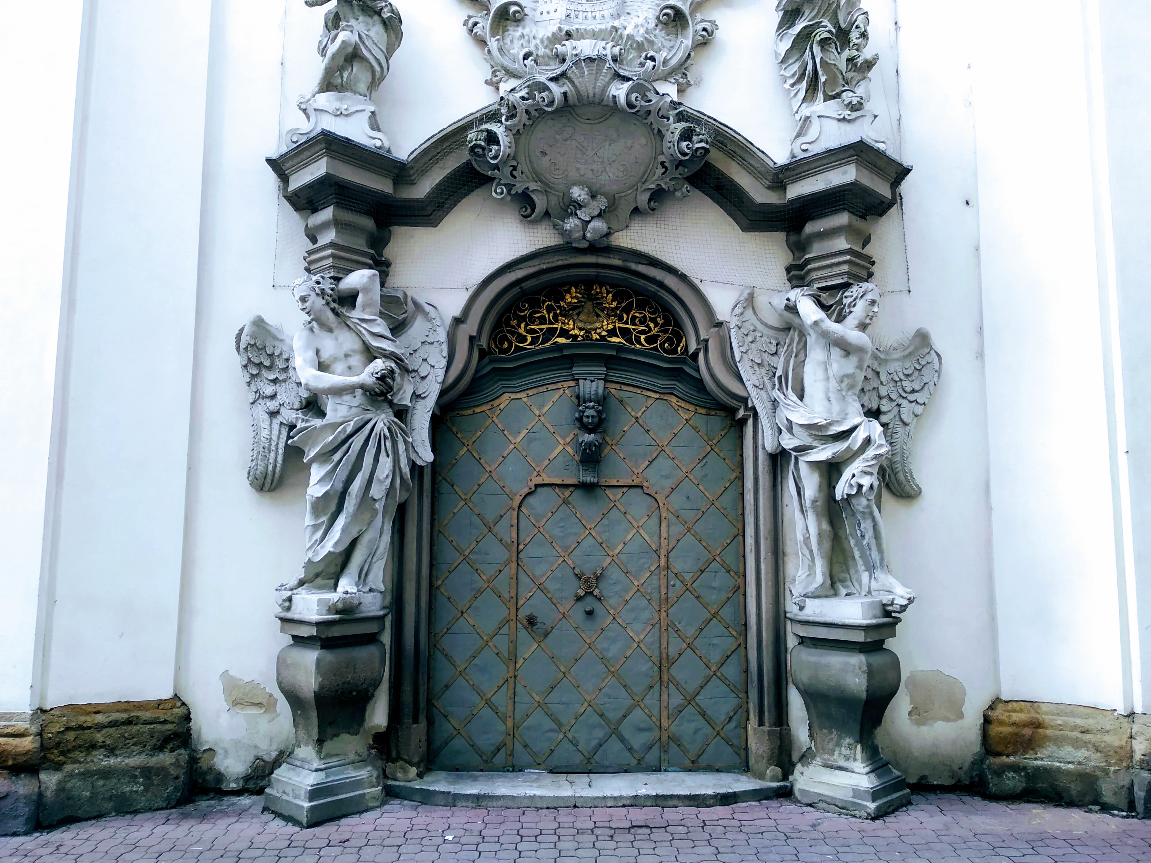 A white wall with an ornate blue door with diamond gilding in an arch held up by statues of angels.