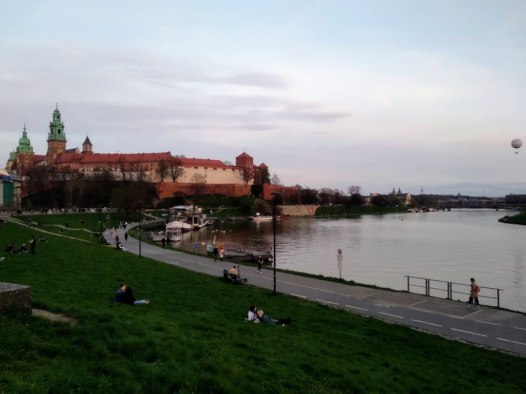 A view of the Wawel Royal Castle and river from a nearby park, in which young couples lie in the grass.