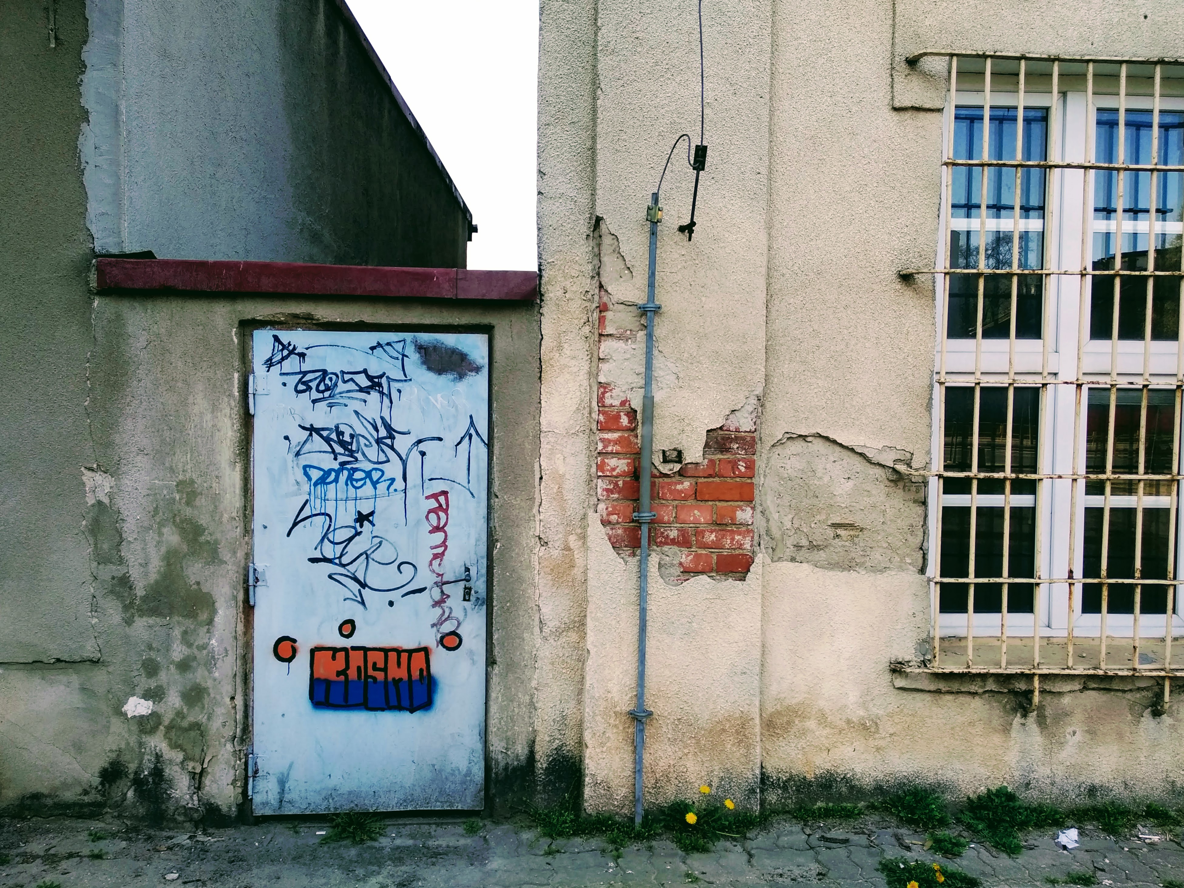 A white metal door between two ageing buildings. The tag Kosmo appears at the bottom in the colours of Ukraine.