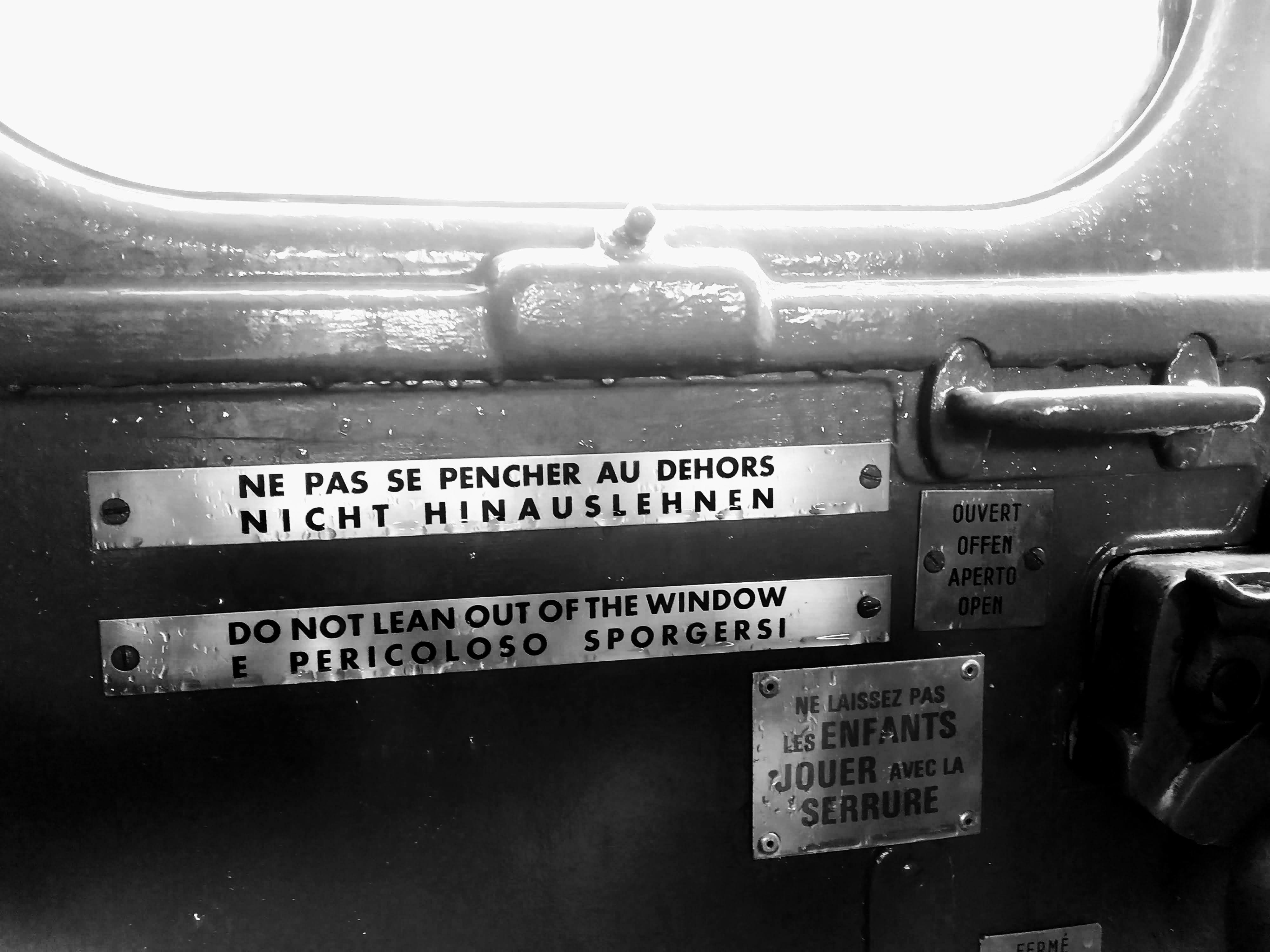 The interior of a wet door in a third-class compartment on a heritage train. "Do not lean out of the window" is written in English, French, German, and Italian as well as a sign in French warning against letting children play with the latch.