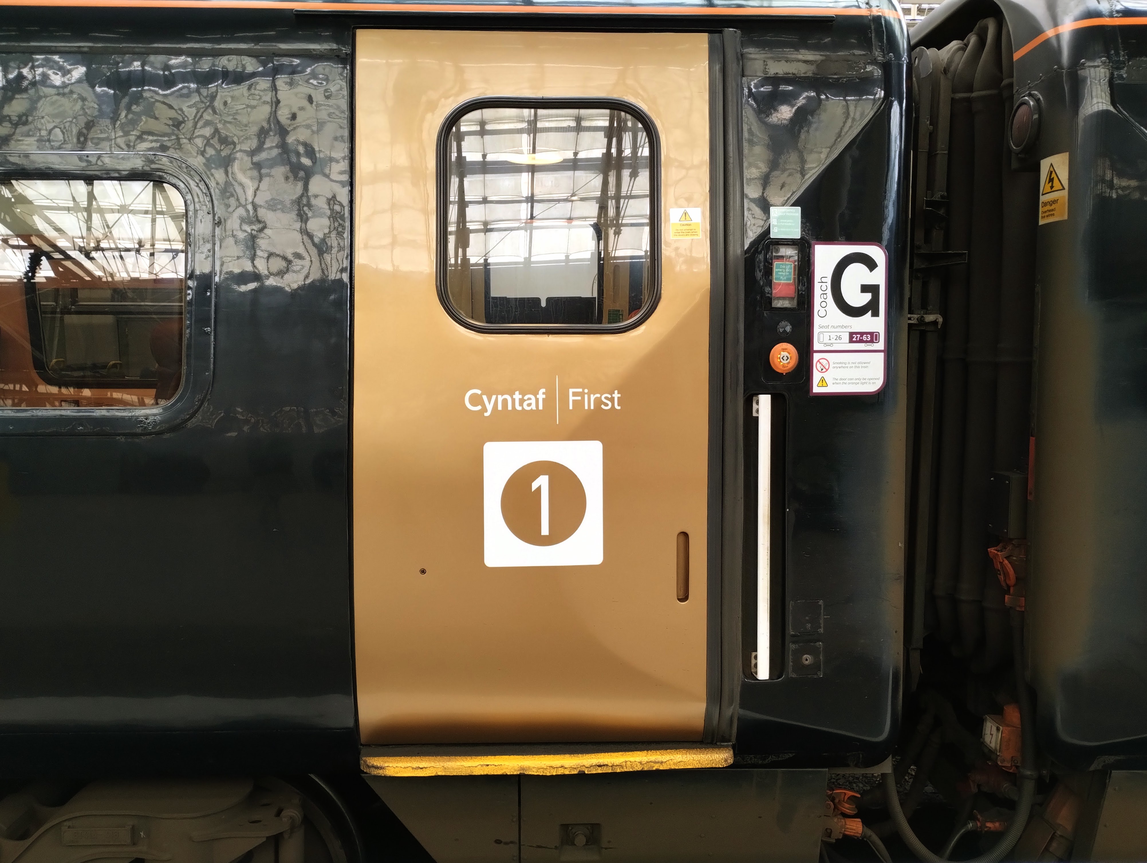 Photograph of a door to first class coach G on a TfW Premium Service. The door is gold, and "Cyntaf/First" is written on it in white letter.