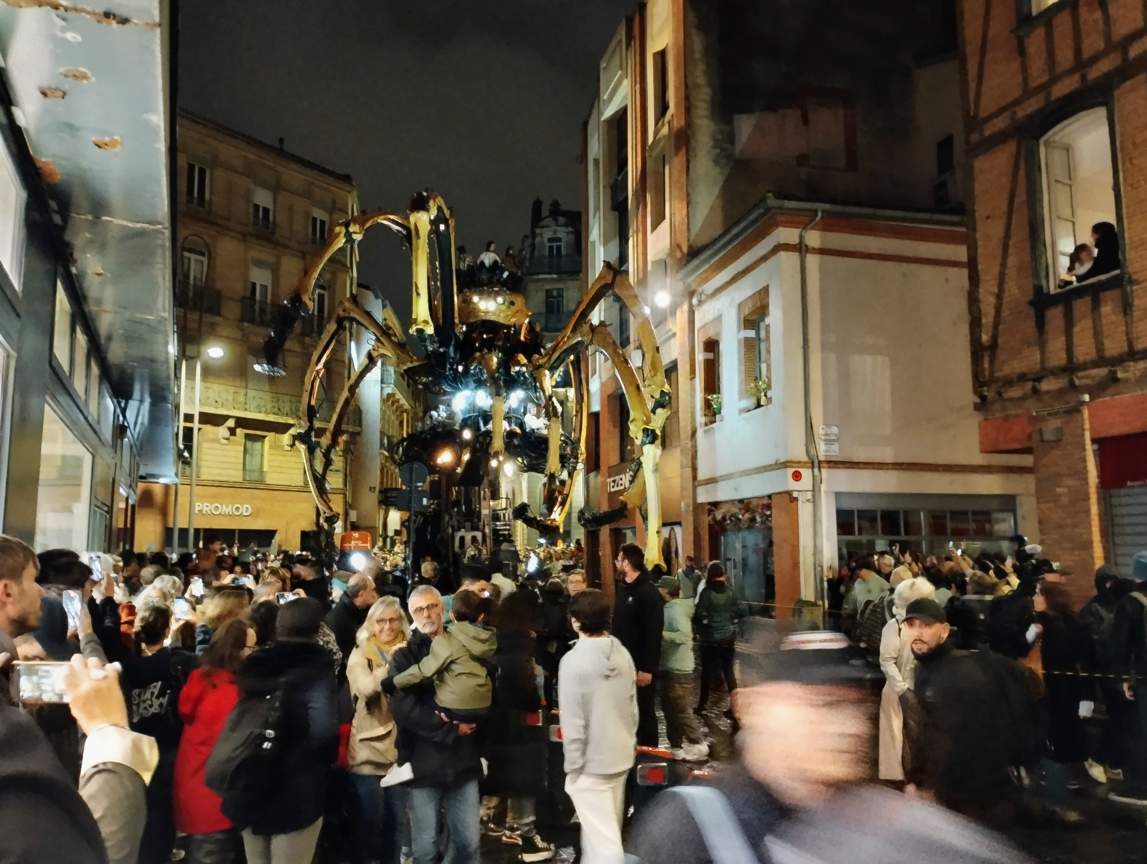 Crowds move out of the way to let a giant mechanical spider through a narrow residential street.
