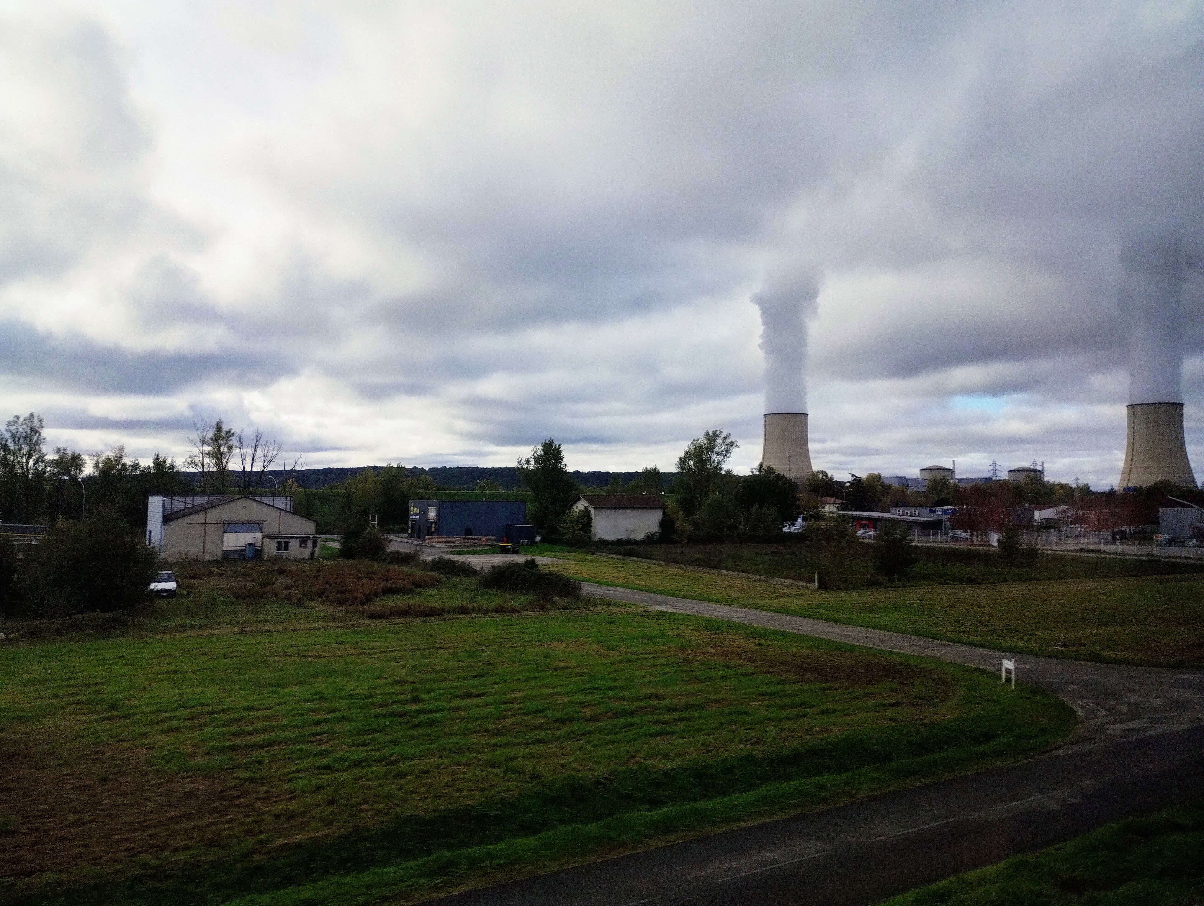 Two cooling towers spewing steam into the cloudy sky dominate a landscape of fields and buildings.
