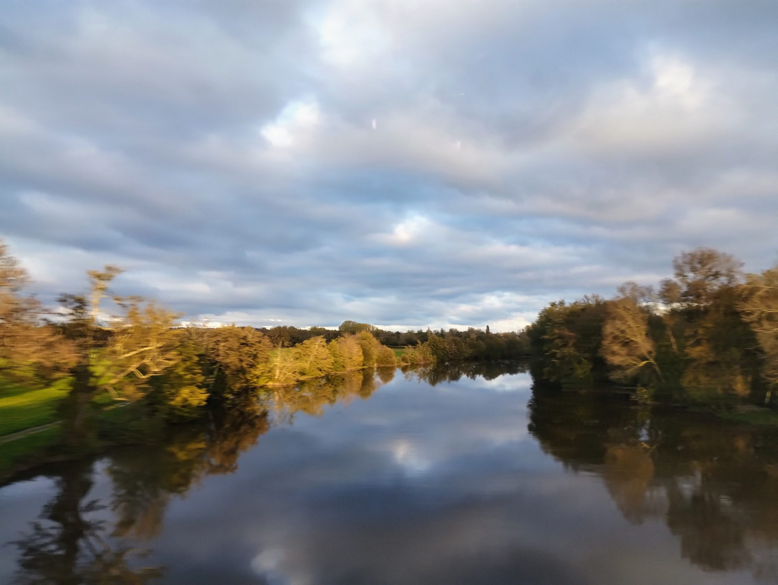 A cloudy sky reflects in a river flanked by trees as seen from a train crossing a bridge.