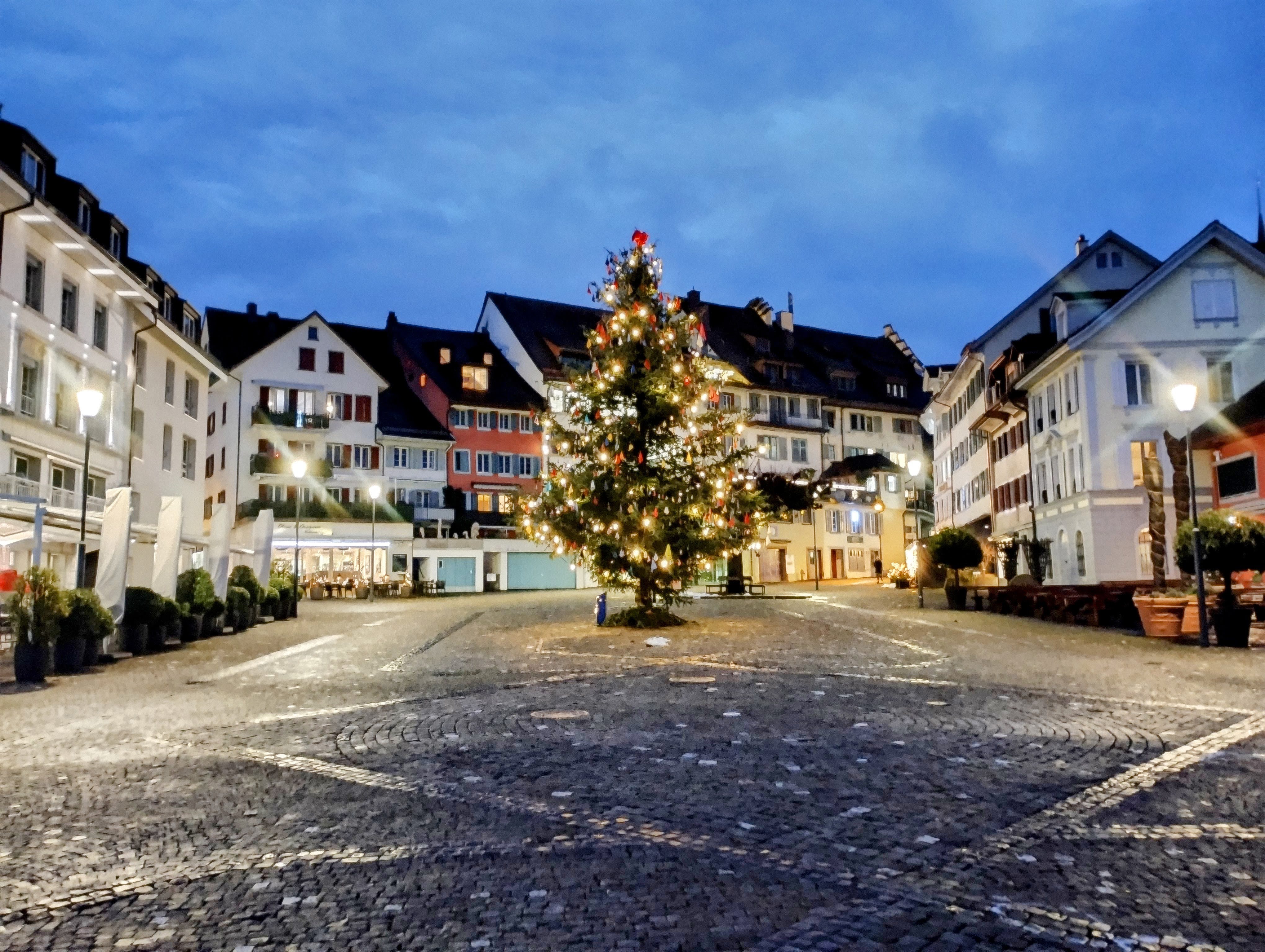 A decorated Christmas tree stands in the middle of a cobbled square, surrounded by white buildings.