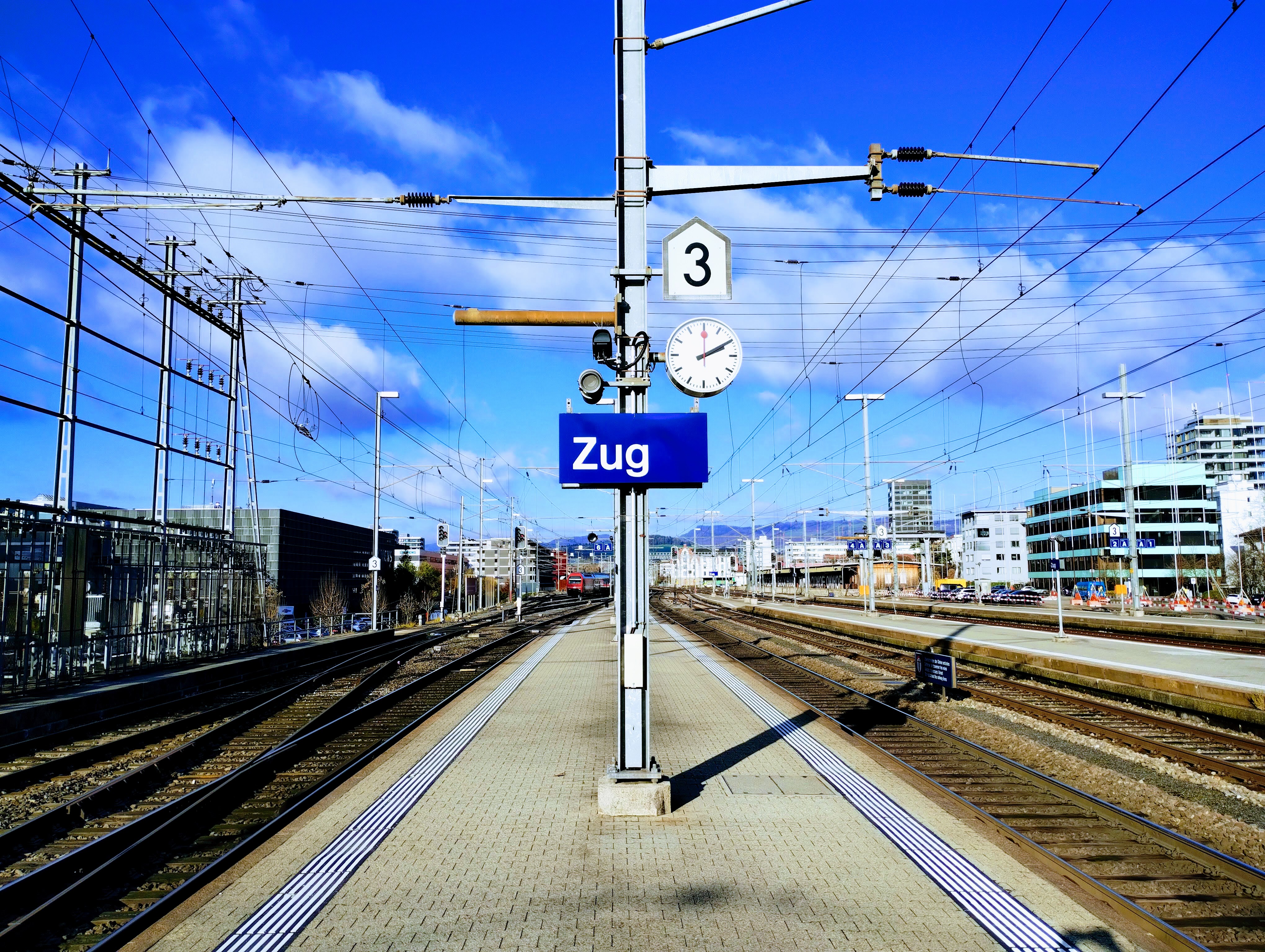 A blue station sign on a platform in Zug, Switzerland. Above the sign, an SBB station clock reads 14:10 as the second-hand pauses for two seconds.