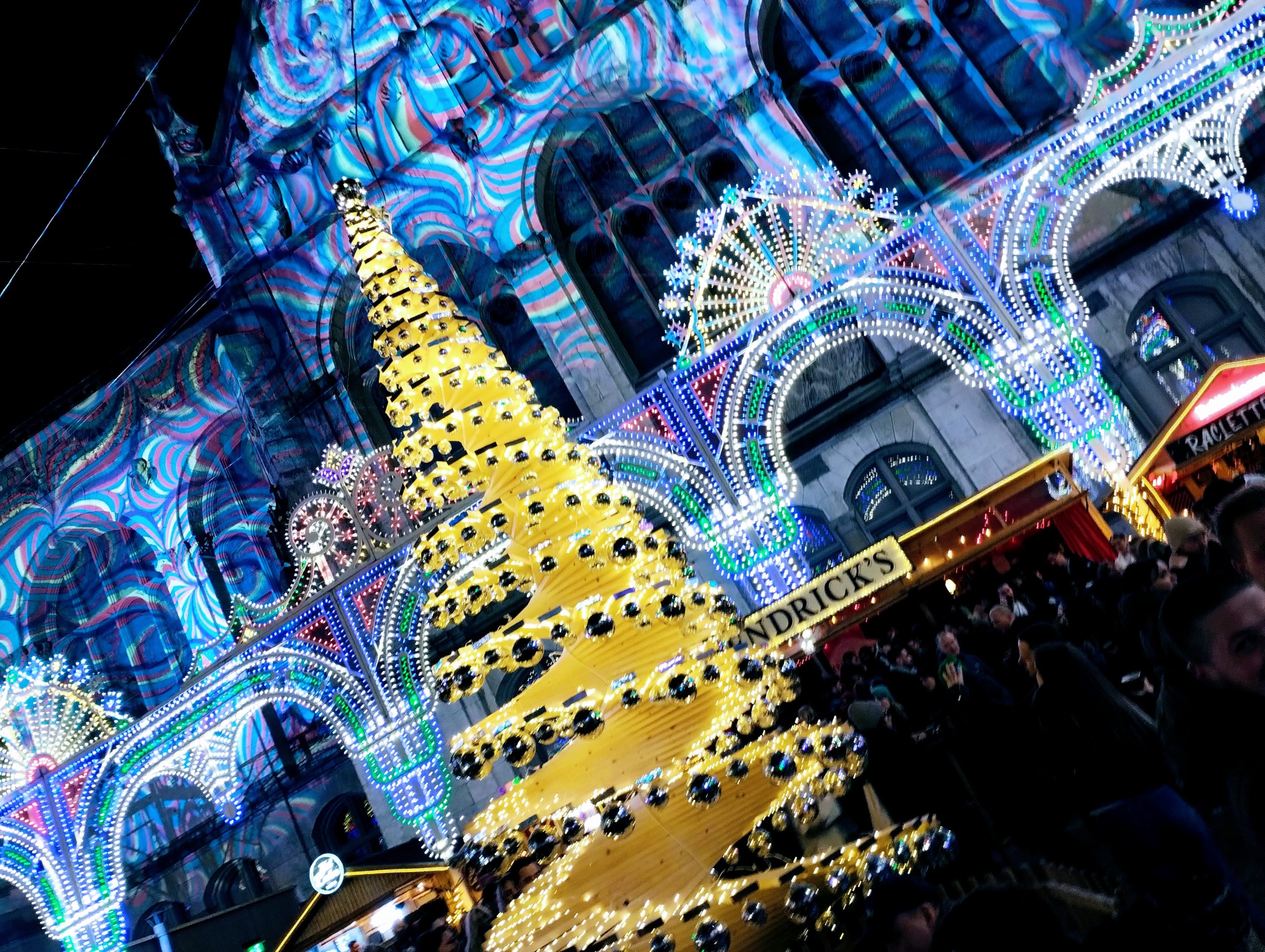 A cream illuminated Christmas tree in front of a multi-coloured light display projected onto a building.
