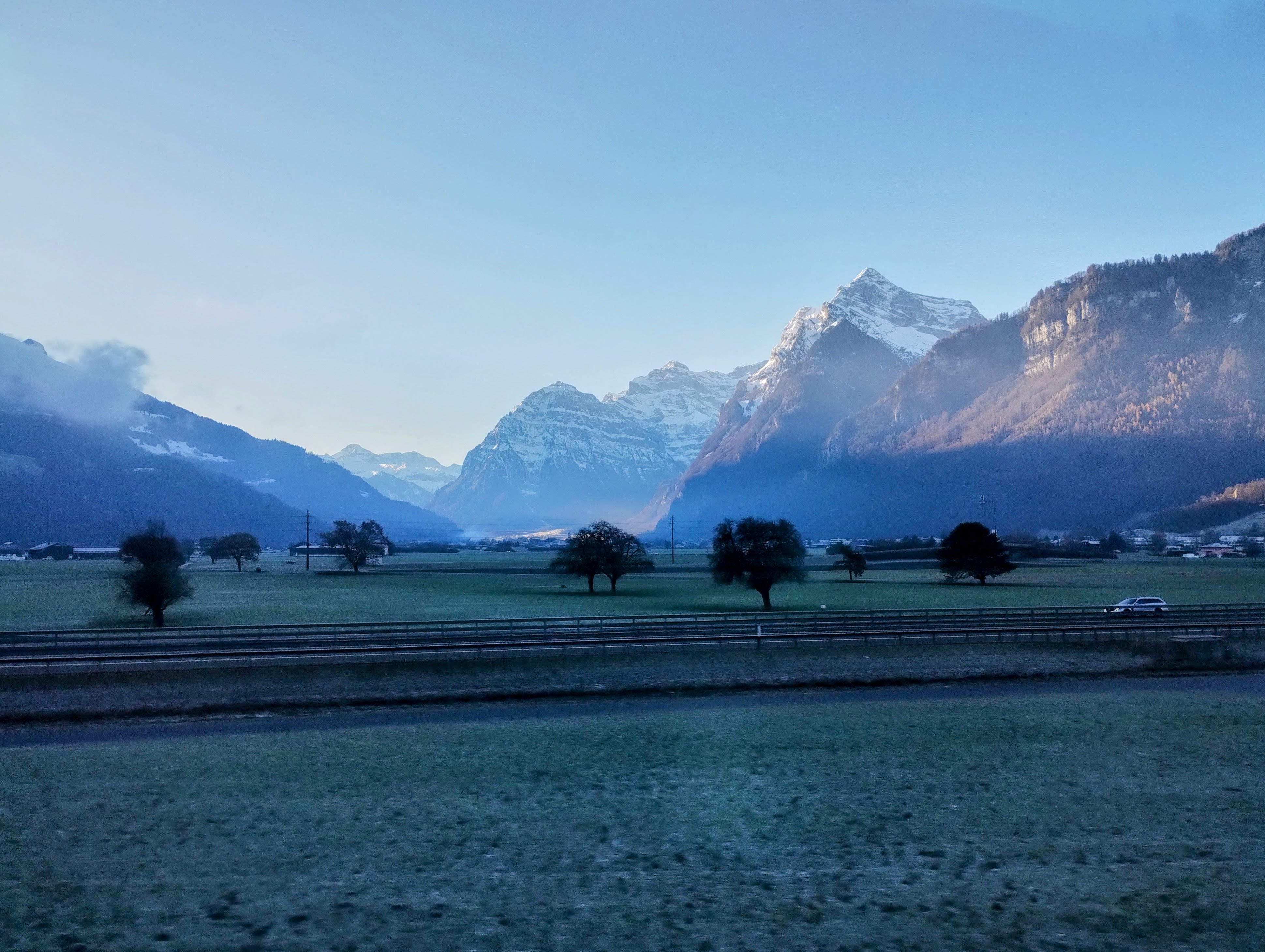 Frosty fields and trees are overshadowed by snow-capped mountains partially illuminated by early-morning sunshine.