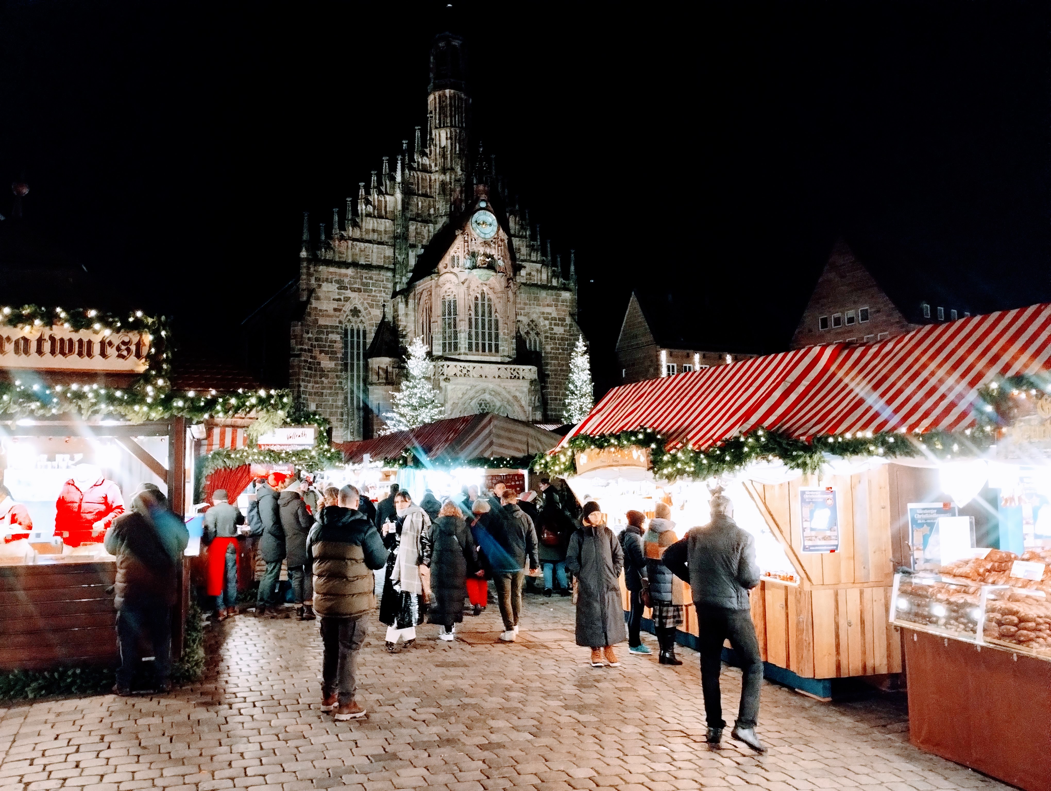 A busy Christmas market with stalls selling Bratwurst and other local treats in front of a gothic church.