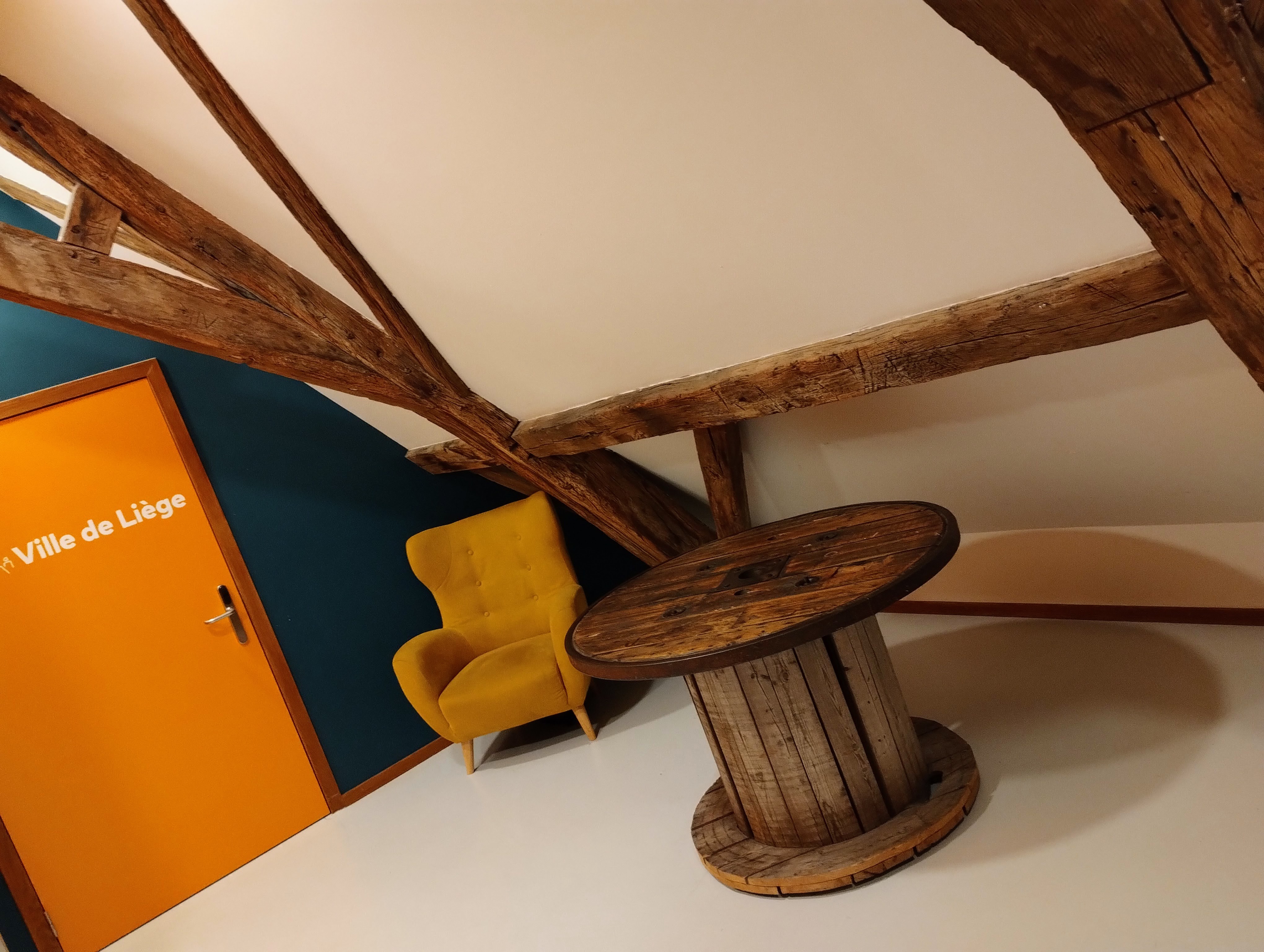 A hallway in a hostel under wooden beams. A wooden cable roll serves as a table to a mustard armchair, next to a plain orange door with the words "Ville de Liège" written on it in white text.