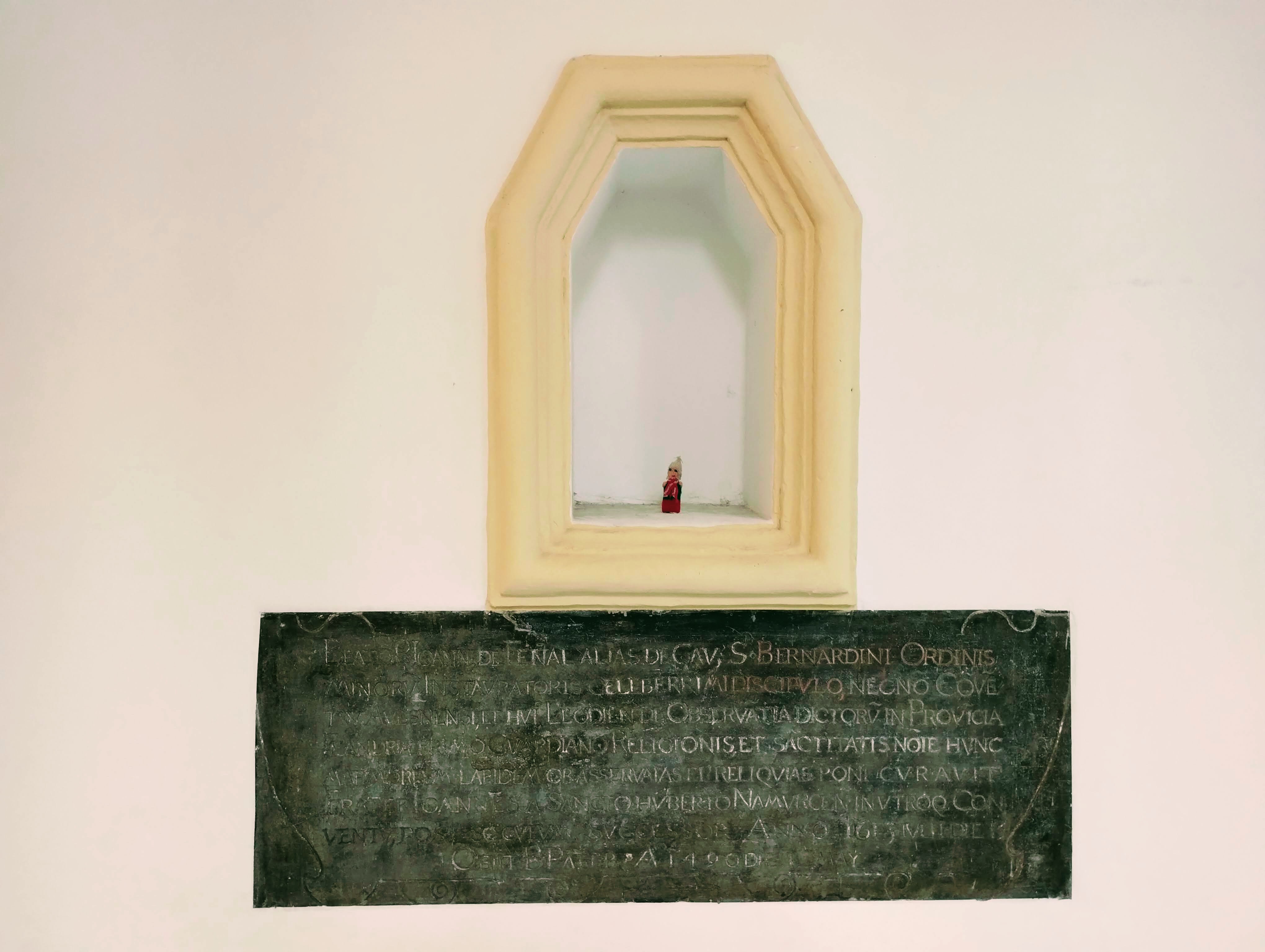 A finger puppet stands in an alcove above a plaque with Latin text which is difficult to read but which ChatGPT suggests "honours a religious figure of the Franciscan (St. Bernardine) order"