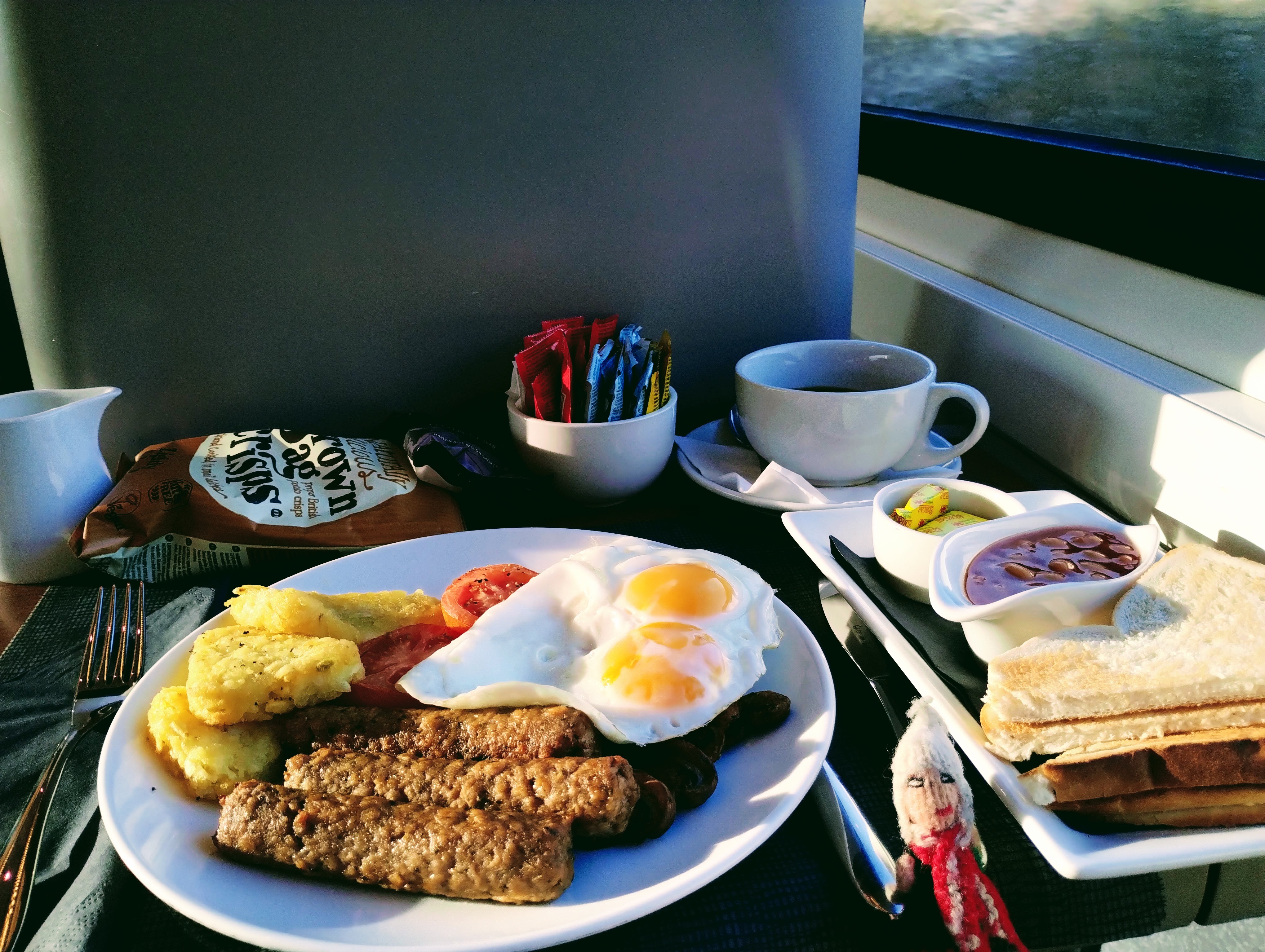 An at-seat cooked breakfast of vegan sausages, hash browns, tomatoes, fried mushrooms, fried eggs, baked beans, toast and butter, and a mug of coffee.