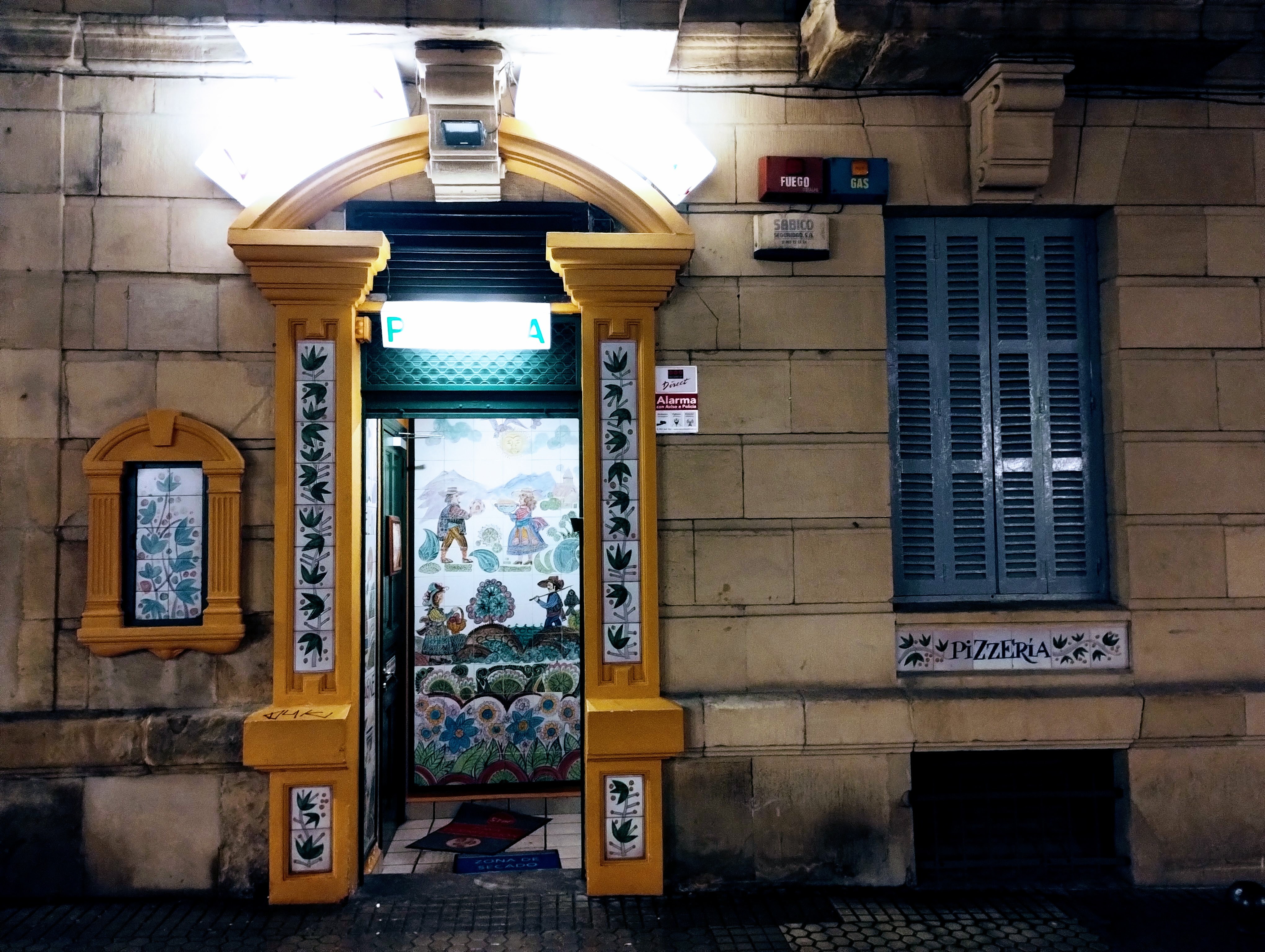 A door to a pizzeria with colourful paintings of people in fields. Around the door a yellow architrave with tiling.
