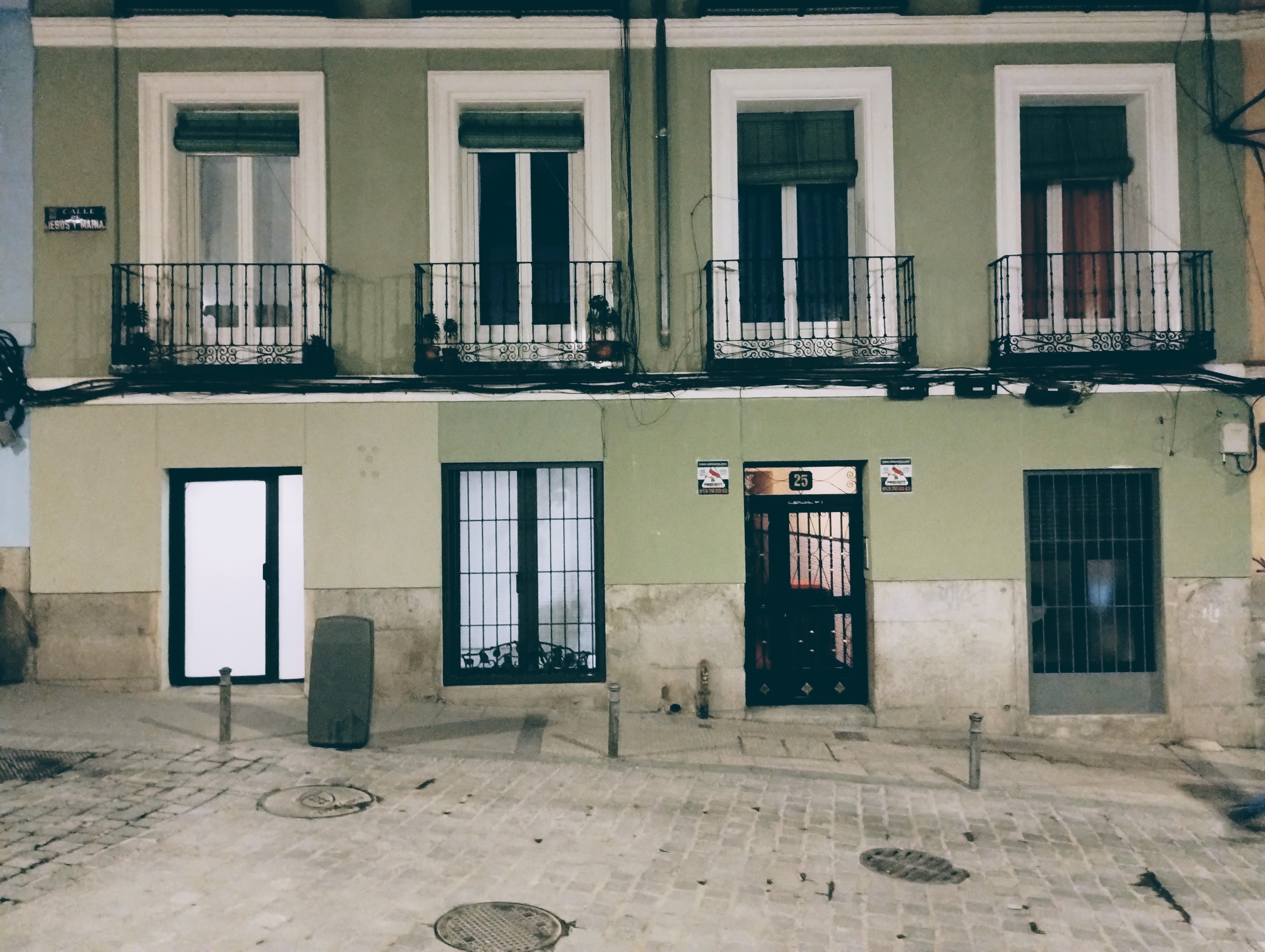 On the ground floor of a sage green residential building, a frosted glass door glows white from the light behind it. On the first floor, four windows with Juliette balconies.