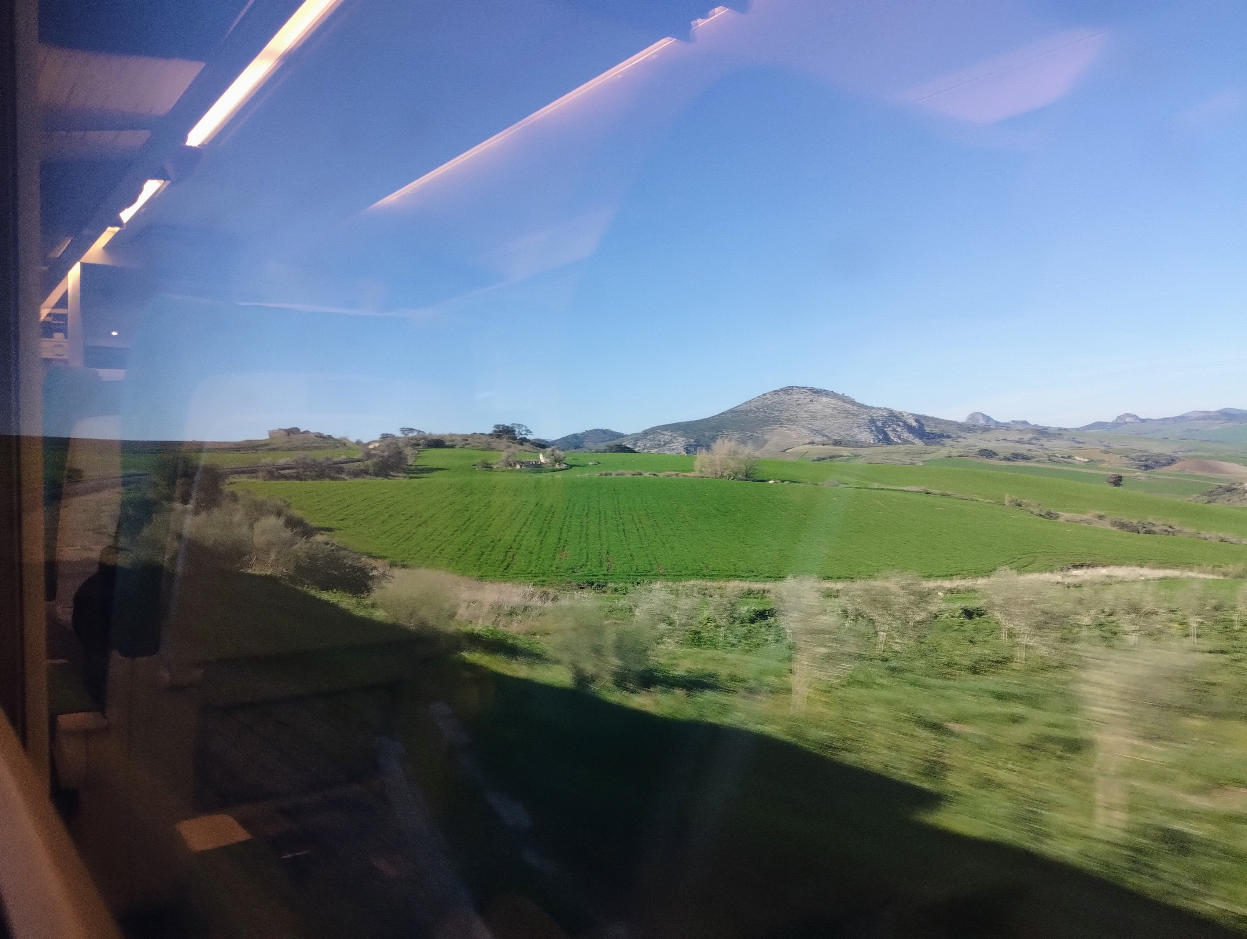 View from a train window, showcasing a lush green field with a rolling hill landscape under a clear blue sky, with the train's interior partially reflected in the window.