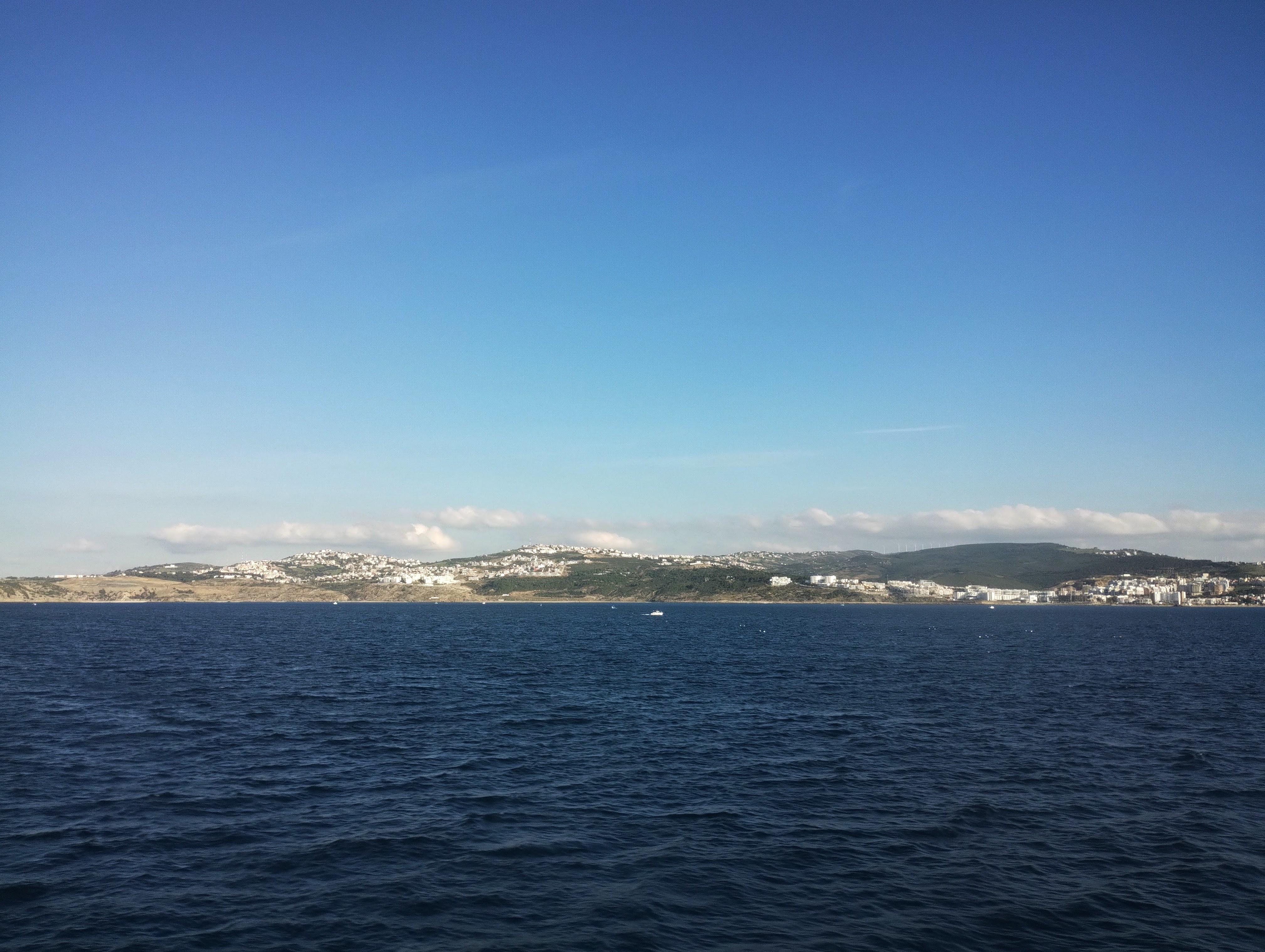 View across a deep blue sea towards the coastline of Morocco with buildings and low hills under a bright blue sky.