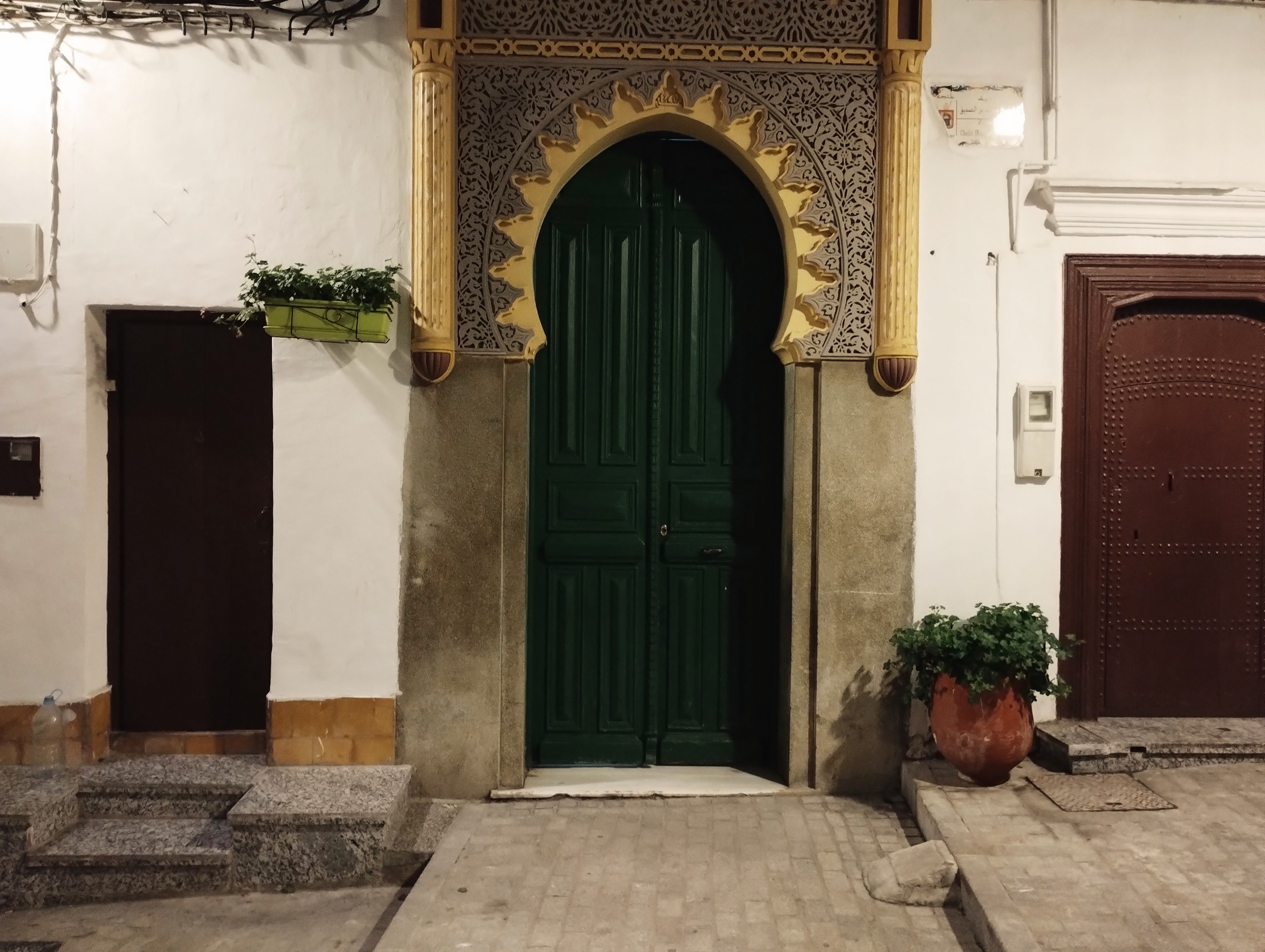 Ornate green wooden door with arched entryway, flanked by decorative pillars with intricate tilework in a white wall, set against a cobblestone and brick street.