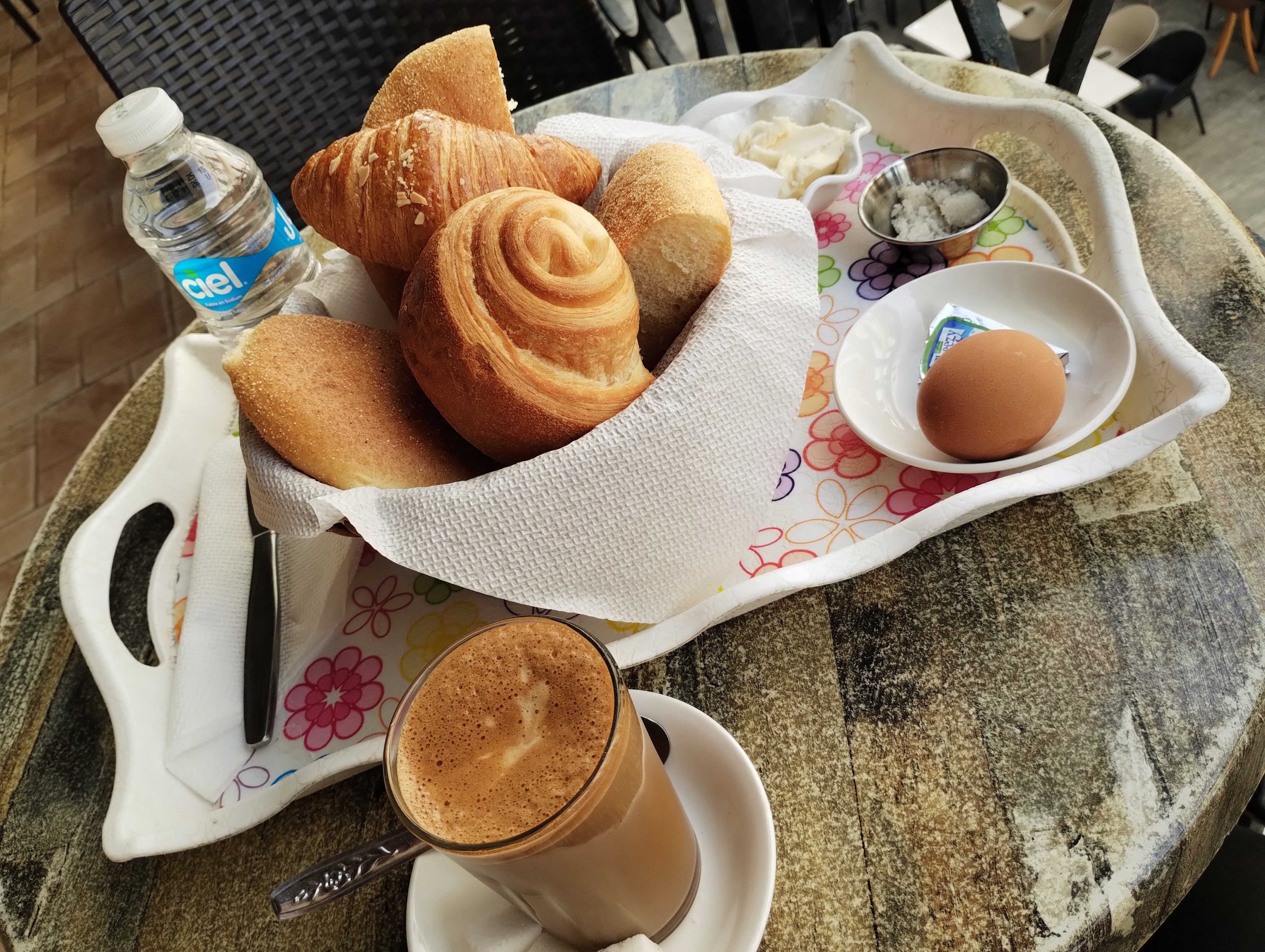Overhead view of a breakfast setting on a patterned tray, including a basket of assorted breads and pastries, butter, salt, a boiled egg, and a bottle of water, next to a glass of coffee on a saucer, all on a weathered, circular table.