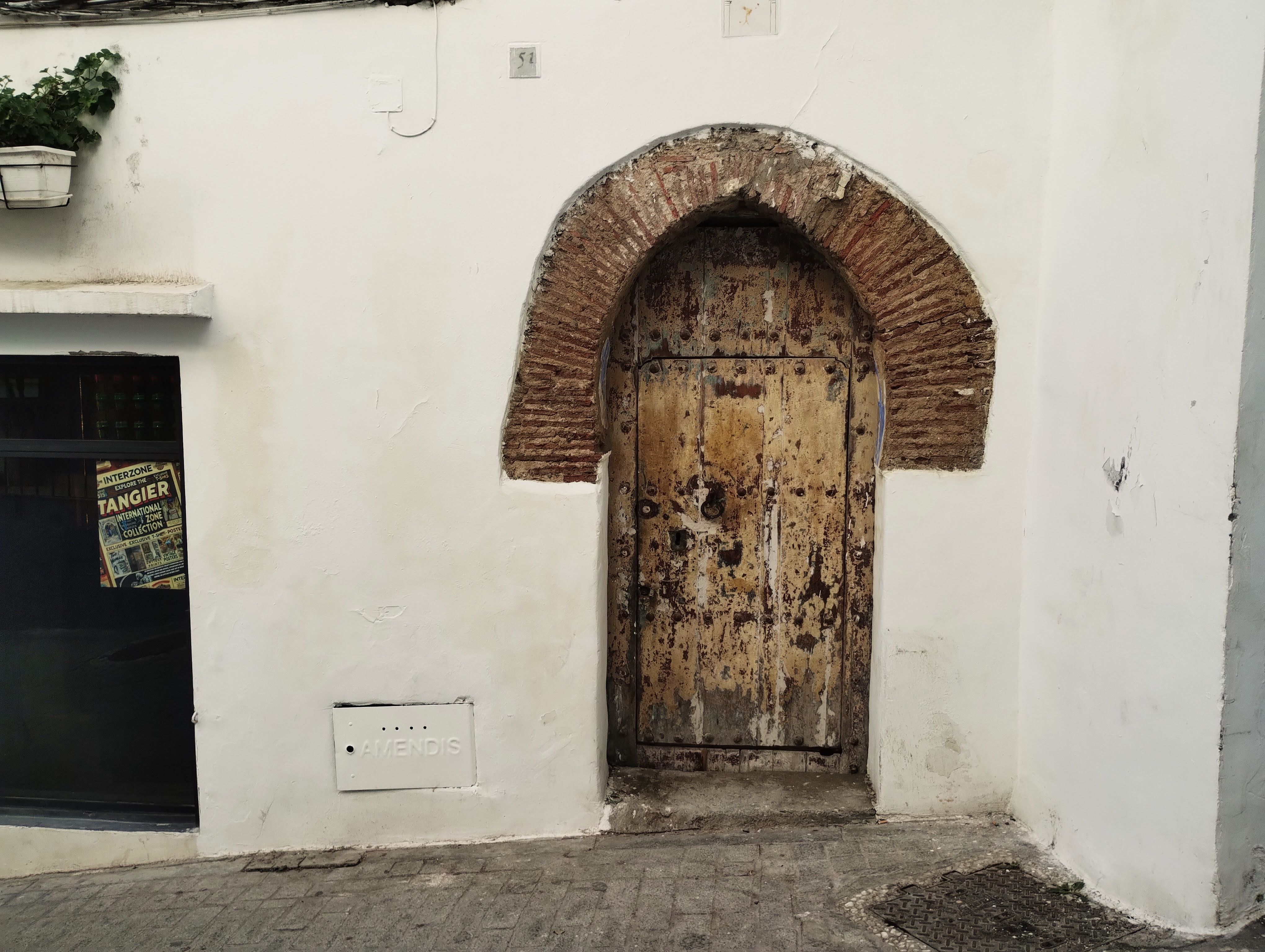 Weathered wooden door in an arched brick frame set in a white stucco wall, adjacent to a street and a building with a poster.