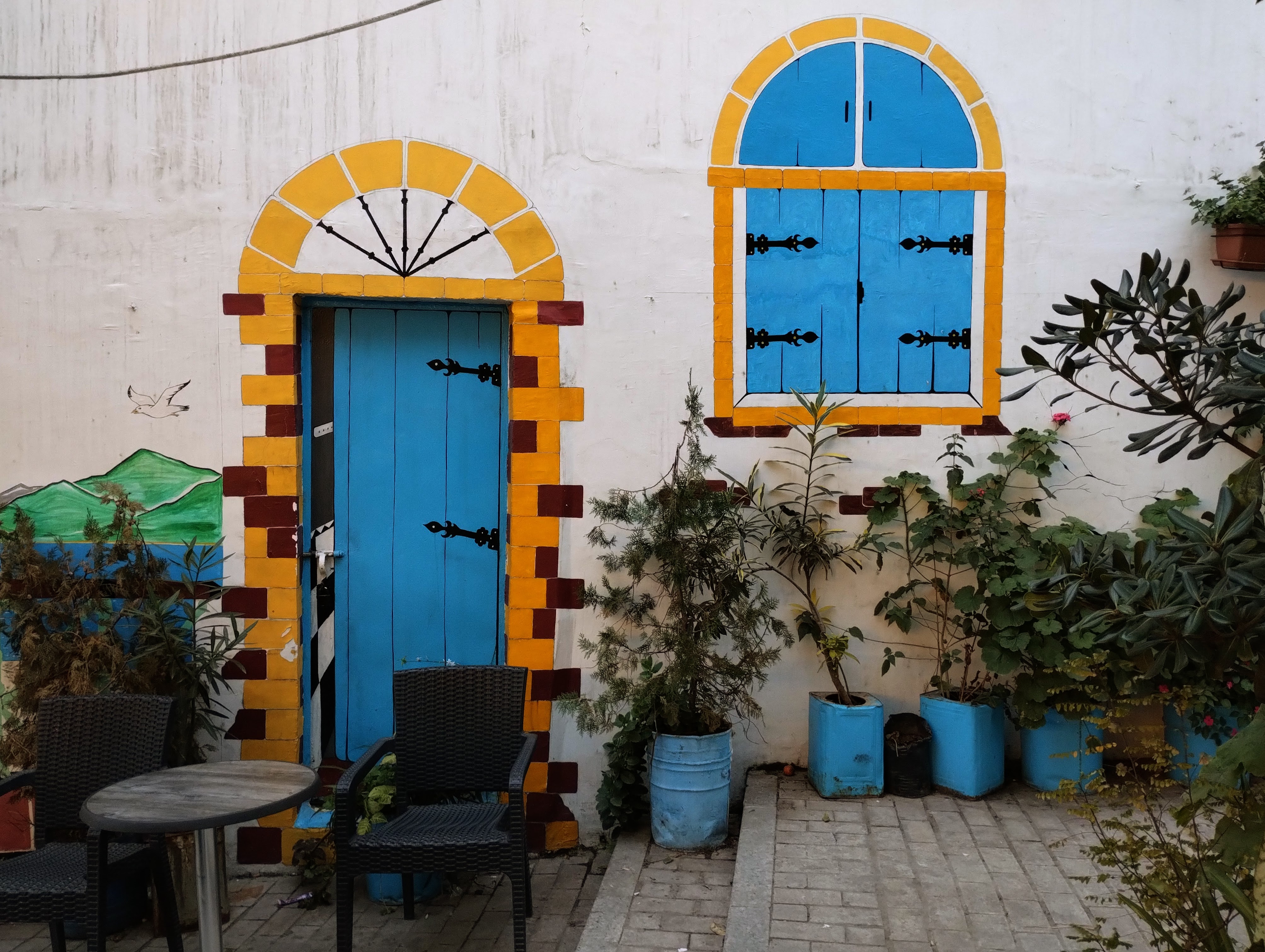 A white painted wall with a blue painted door and window with arched yellow frames, black decorative hinges, and brickwork border, flanked by potted plants and a small table and chairs.